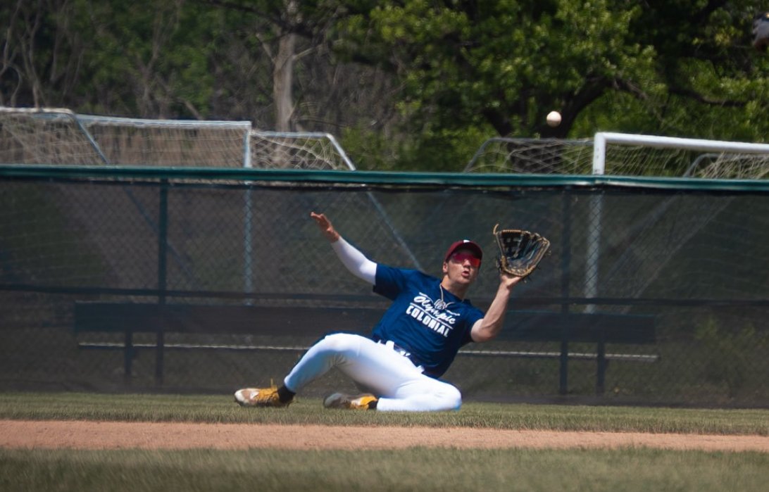 A baseball player is on the ground, reaching with his mitt to catch a ball near a fence on a baseball field.