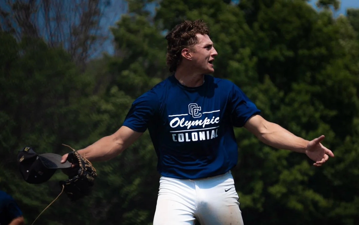 A young male baseball player wearing a blue shirt with 'Olympic Colonial' printed on it, holding a baseball glove and helmet, standing outdoors with trees in the background.