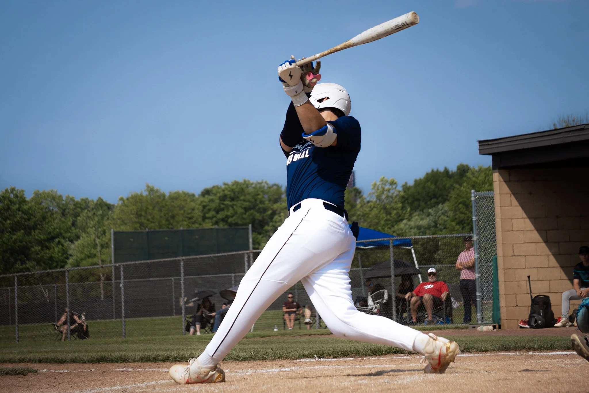 A baseball player swings a bat during a game on a sunny day, with spectators watching in the background.