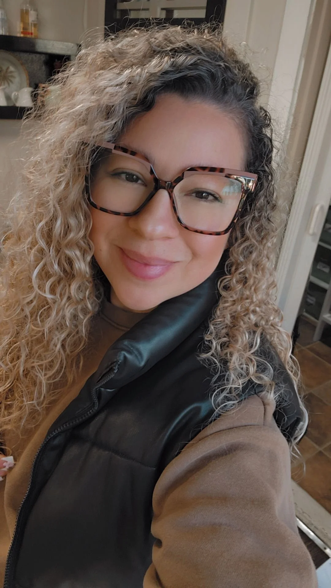 A woman with curly dark hair, glasses, and a beard, sitting indoors with a plant in the background.