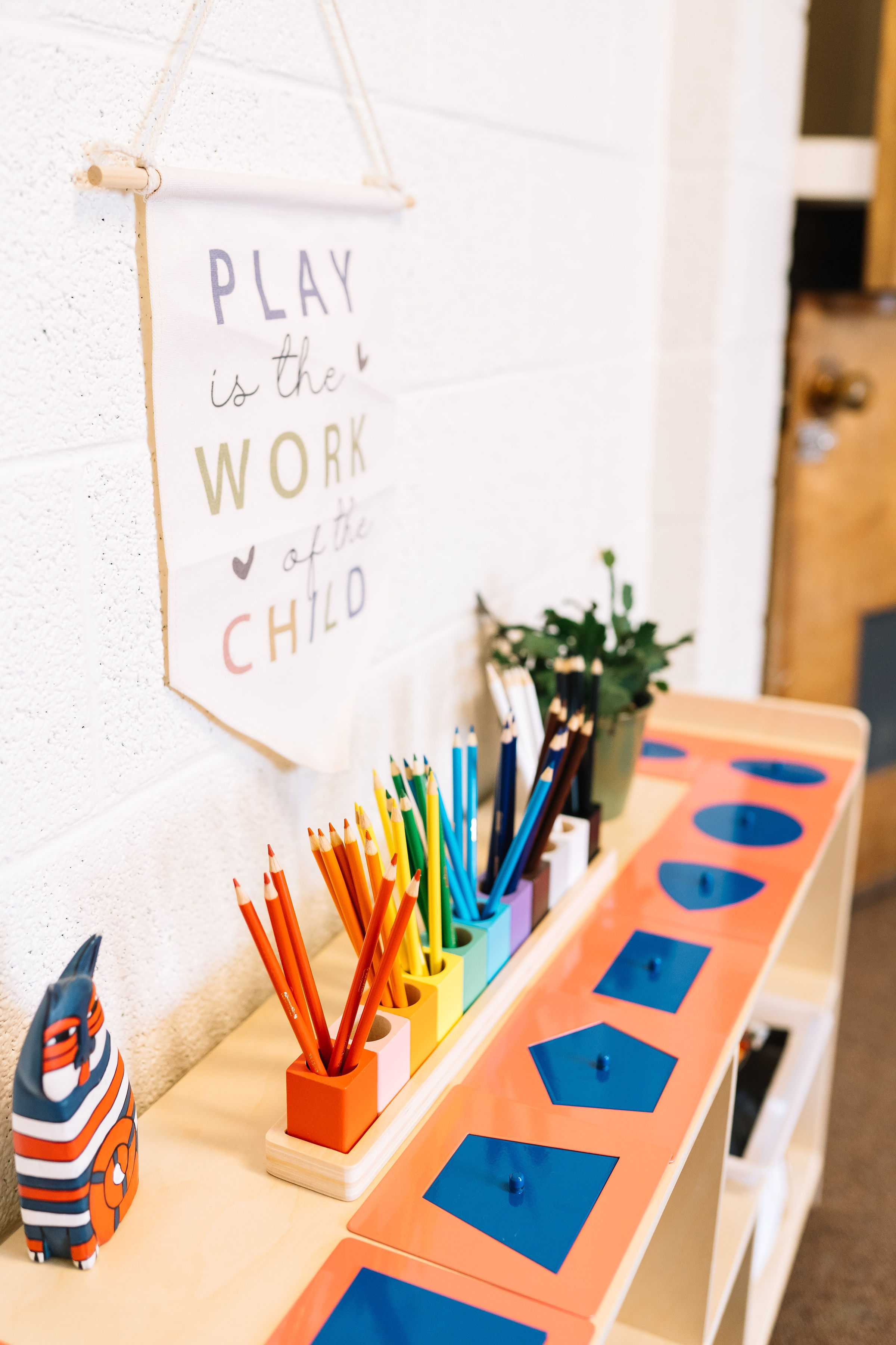 Children's play area with colorful pencils in holders, a potted plant, and a sign on the wall that reads 'Play is the work of a child.'