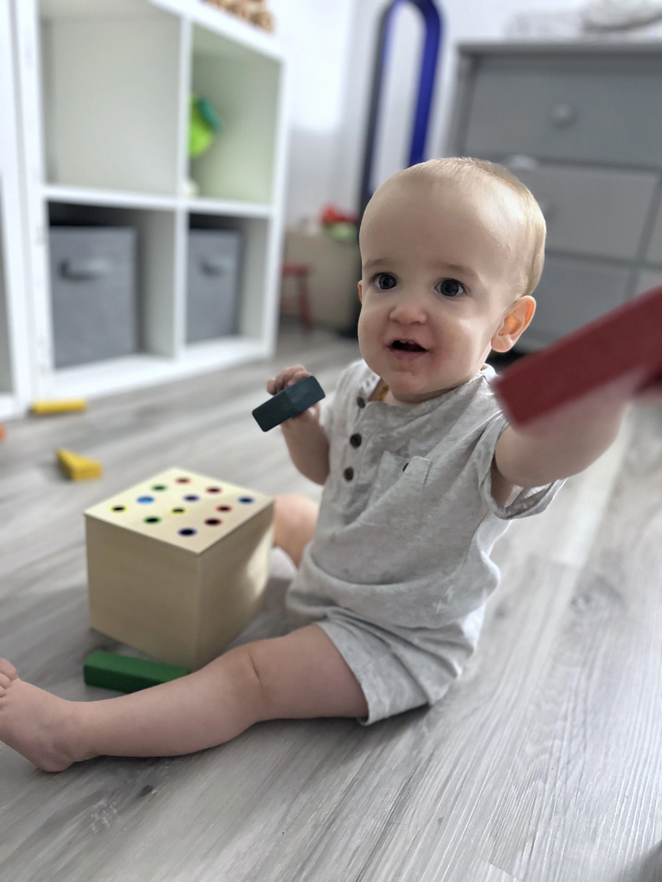 A baby sitting on the floor inside a room, holding a black toy in one hand and reaching out with the other. There are toys and storage bins around, with a white shelving unit and a grey dresser in the background.