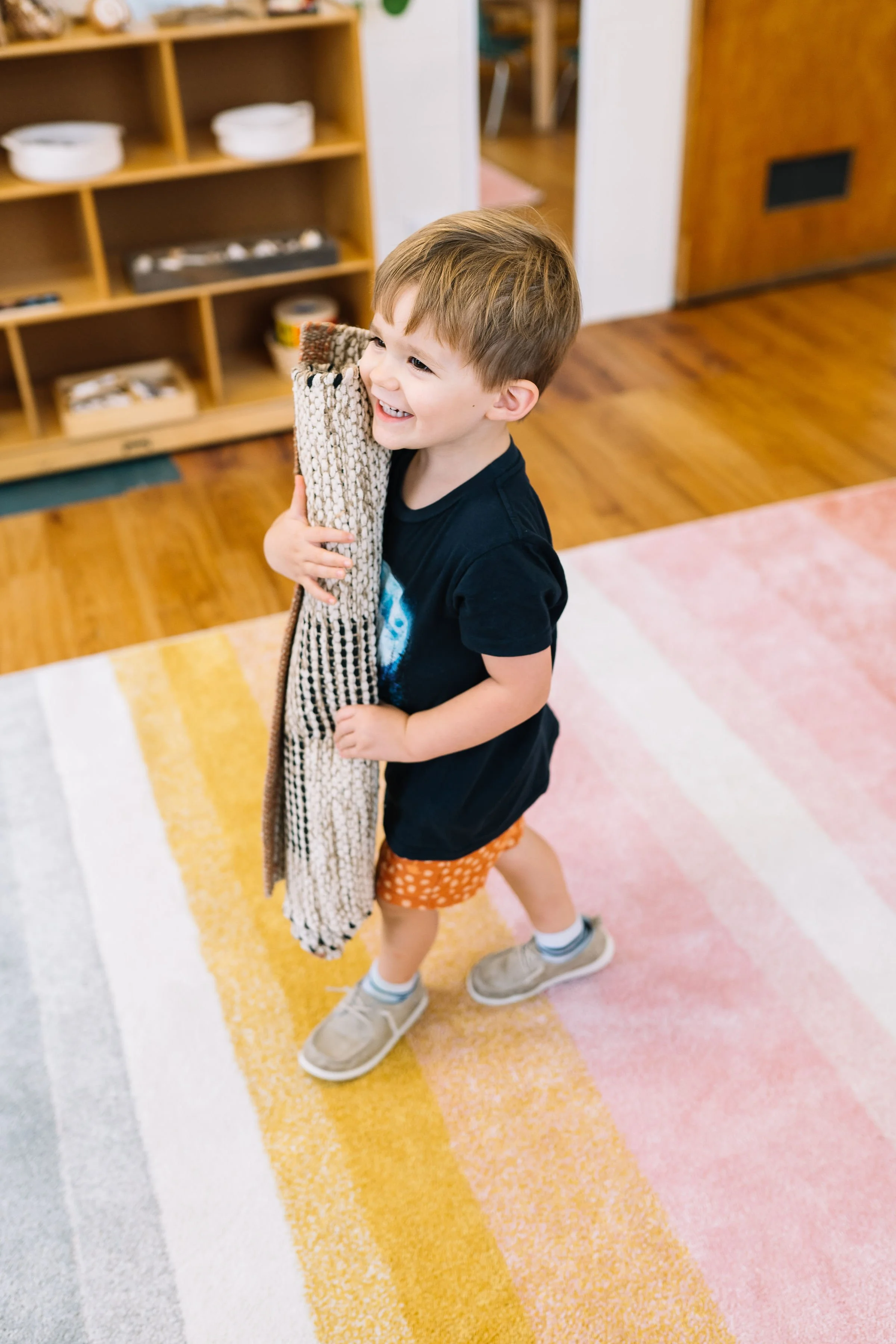 A young boy with short light brown hair, wearing a black t-shirt, orange polka dot shorts, and beige sneakers, smiling and hugging a knitted sock while standing on a colorful striped rug in a room with wooden flooring and a wooden shelf in the background.