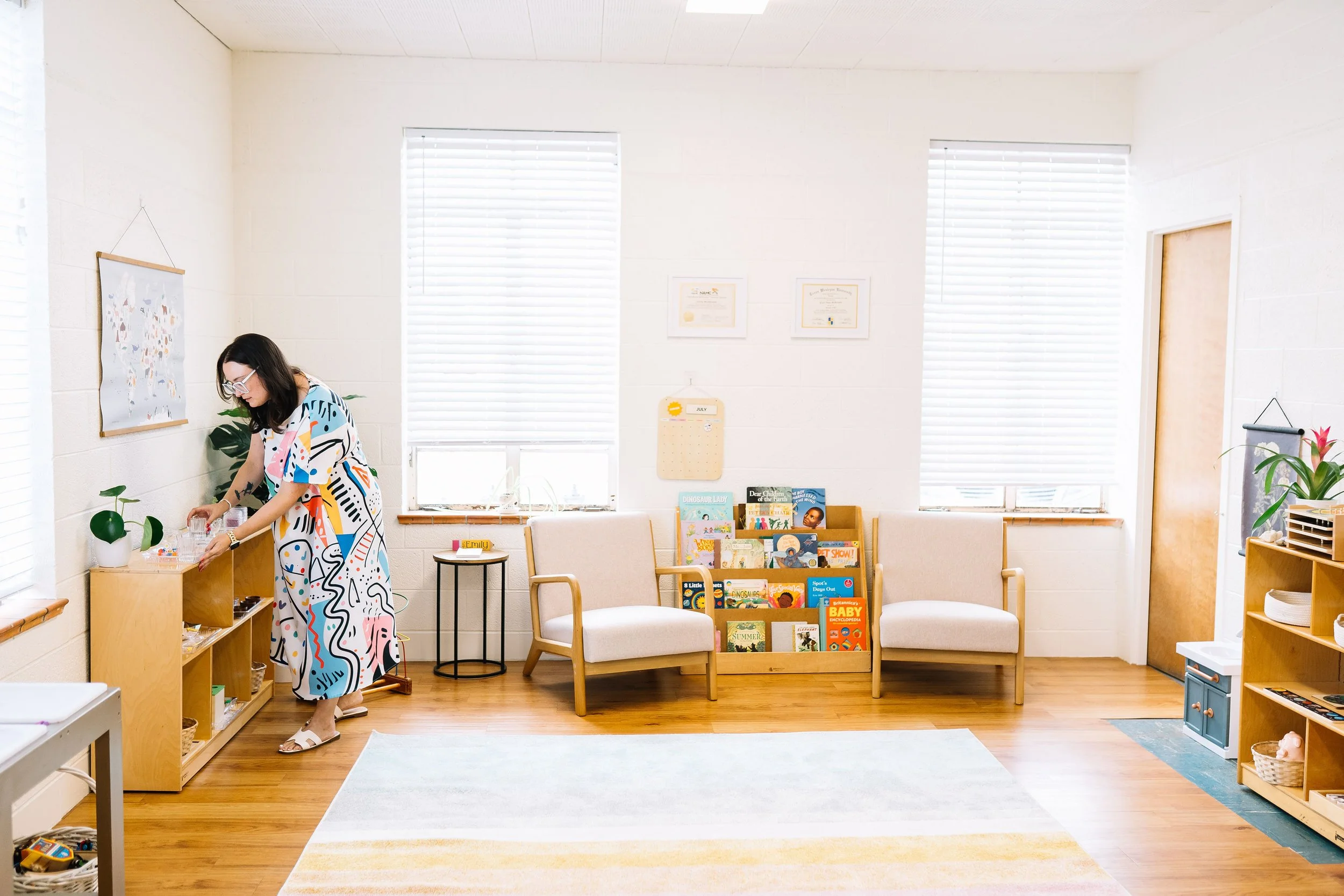 A woman in a colorful dress organizes items on a wooden shelf in a bright, welcoming children's waiting room or classroom, furnished with chairs, a bookshelf, and decor.