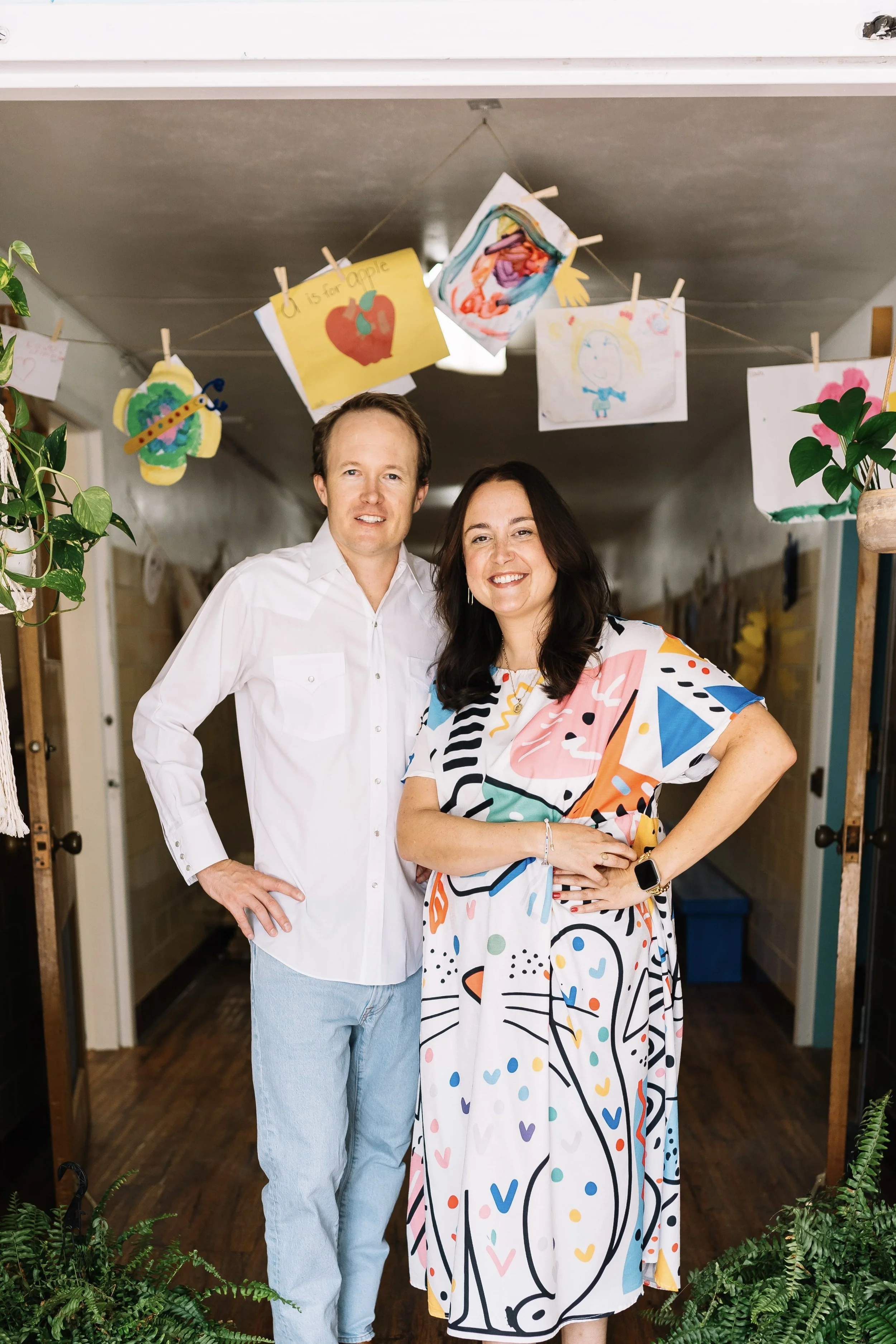 A man and woman standing together indoors, smiling, with children's artwork hanging from the ceiling behind them.