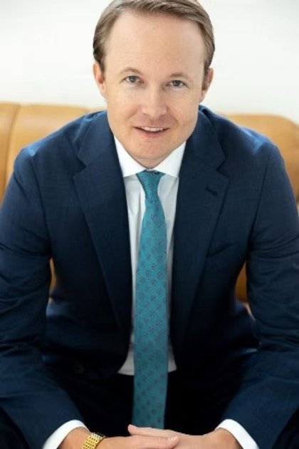 A man in a blue suit and tie sitting in an office environment with a wooden desk and yellow furniture in the background.