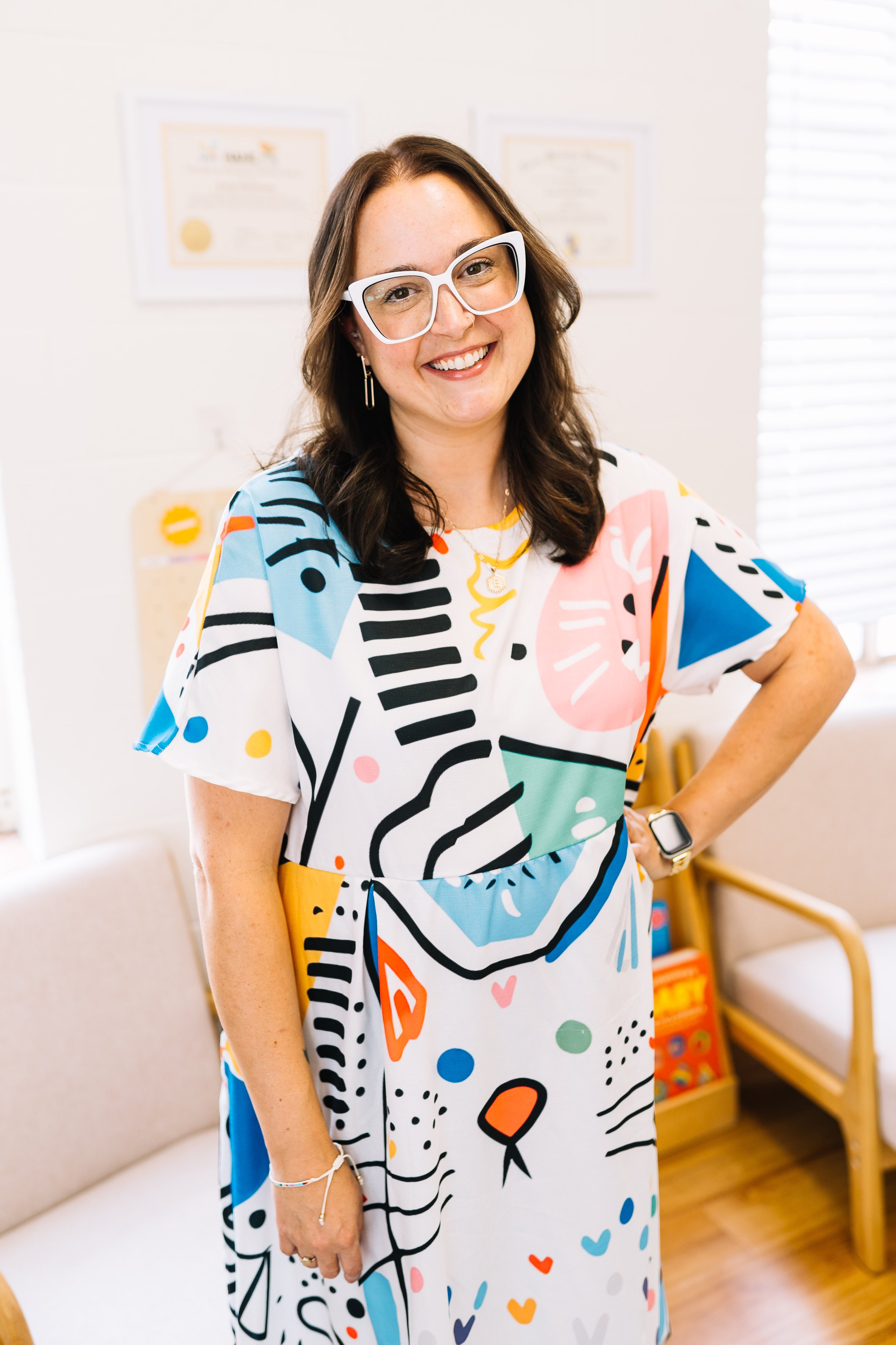 A woman wearing glasses and a colorful abstract patterned dress, standing indoors with a smile, with framed documents on the wall behind her.