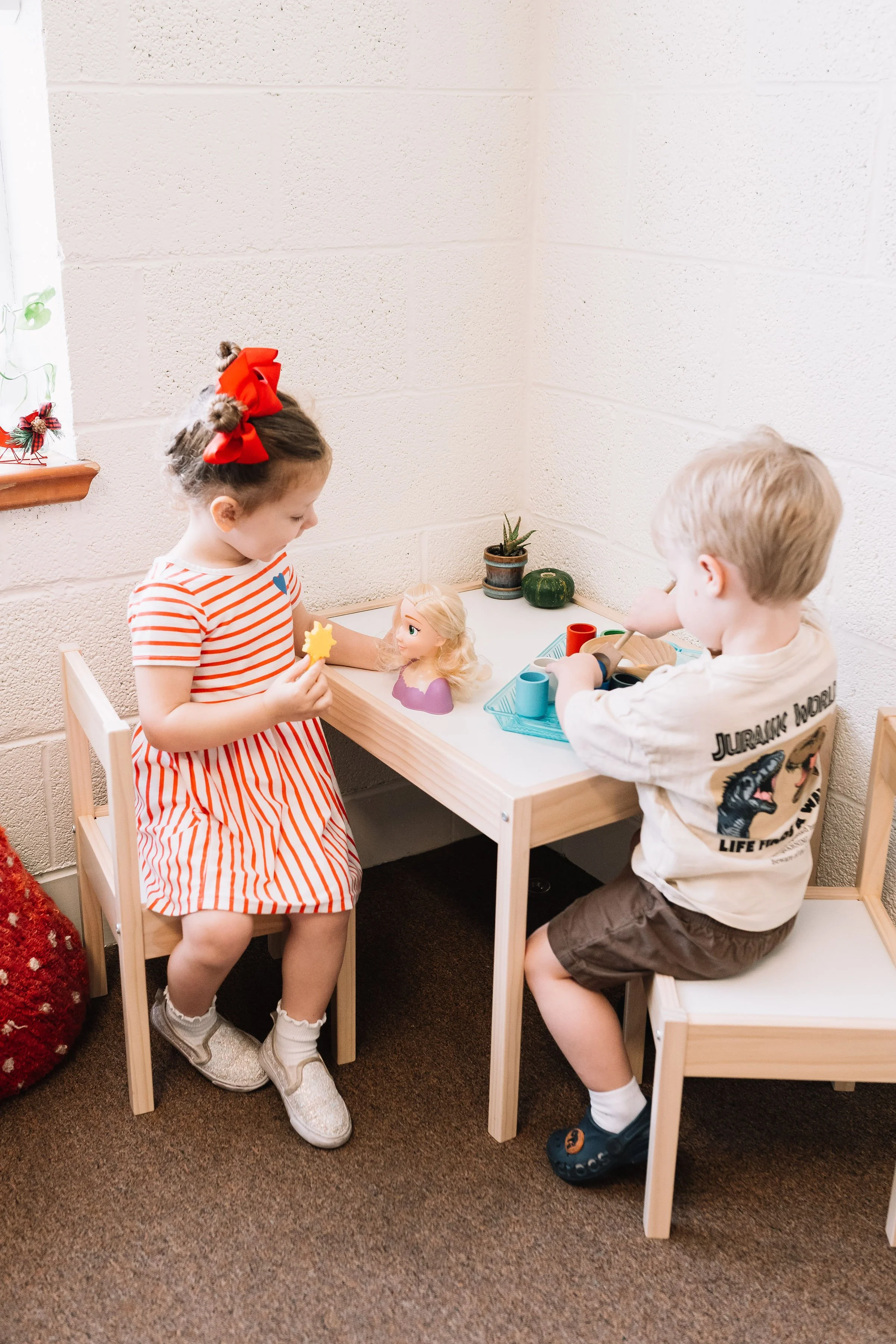 A young girl and boy playing at a small wooden table with toys and a small plant, inside a room with beige brick walls and a window.