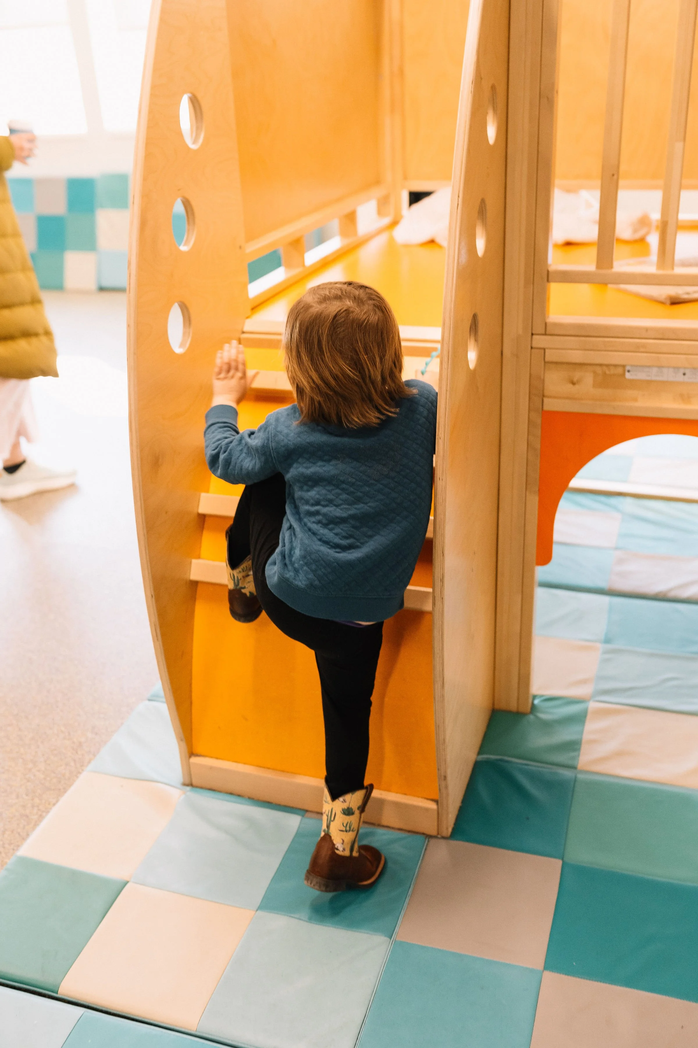 Child climbing stairs inside a wooden play structure in a playroom with colorful padded flooring.