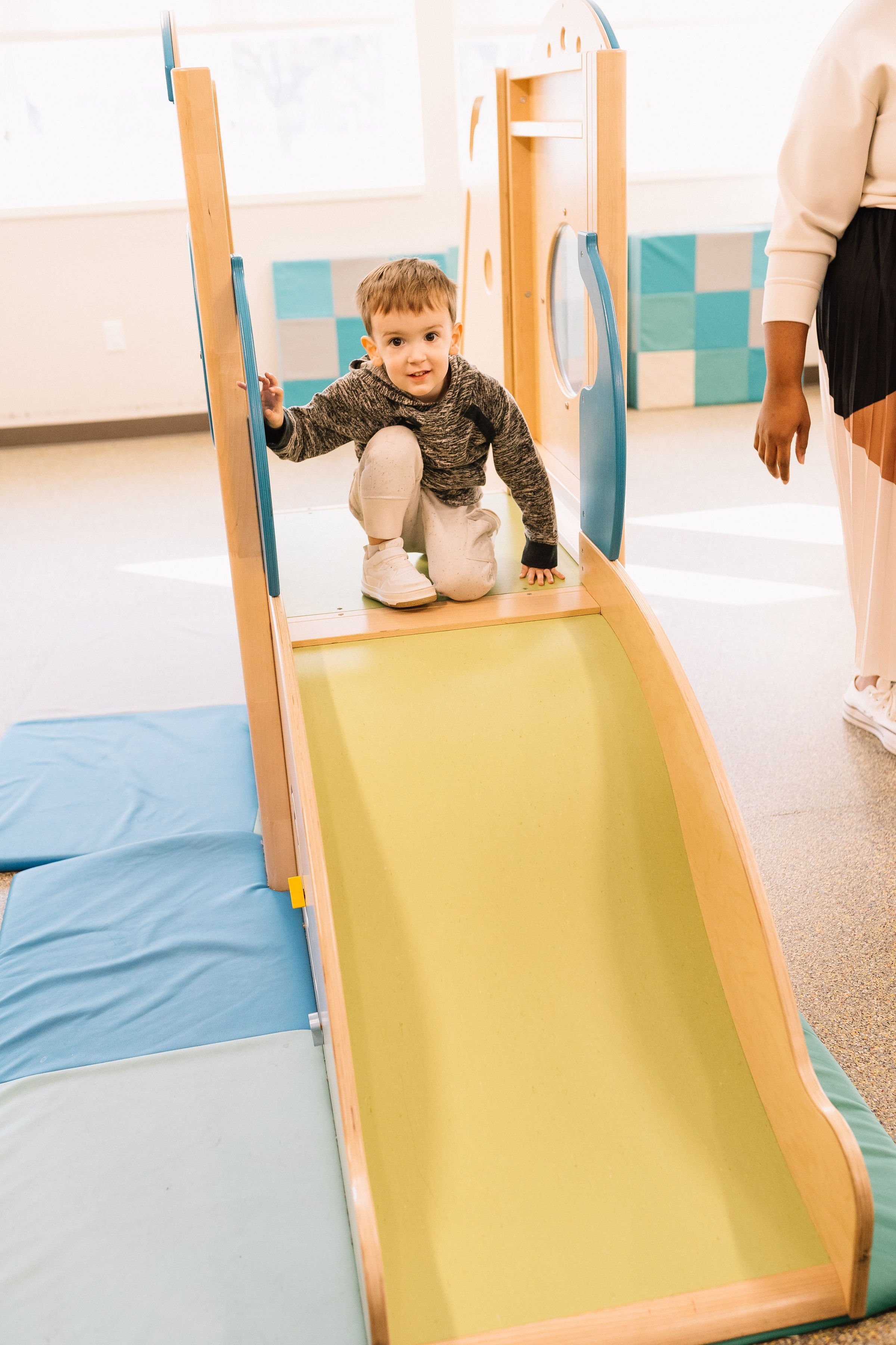 A young boy crouching on a small indoor slide in a playroom, wearing a gray hoodie and beige pants, with an adult partially visible nearby.