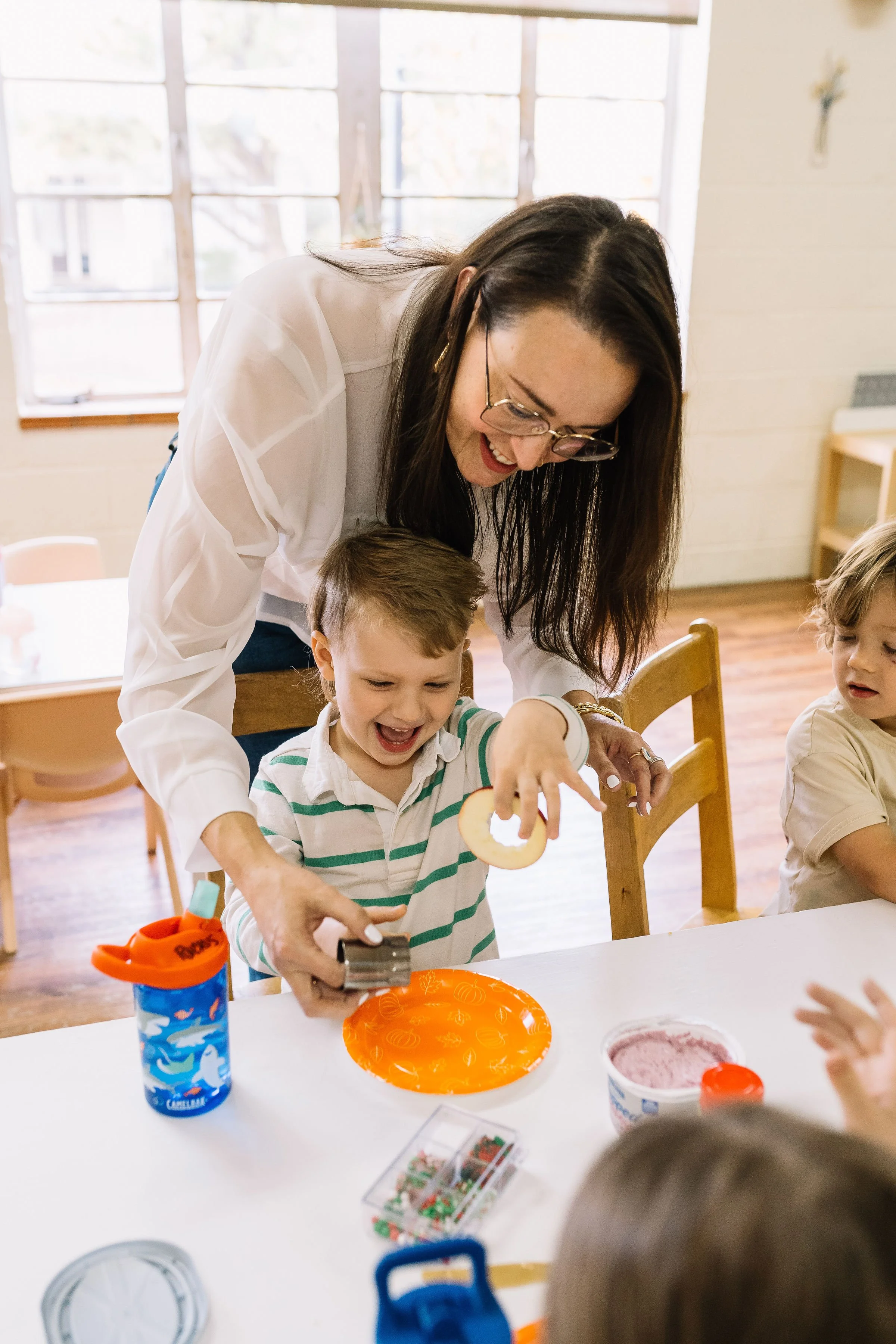 A woman helping a young boy create a craft project at a table during a gathering, with children and craft supplies on the table, in a bright room with large windows.