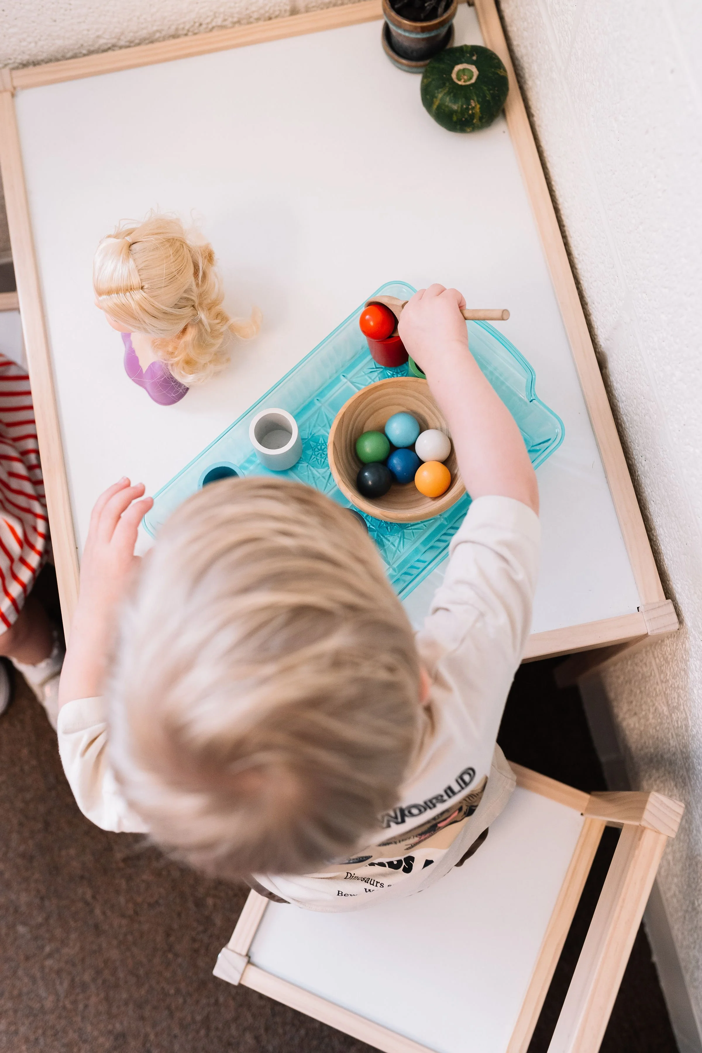 A young child playing with colorful wooden balls and a toy hammer at a small white table.