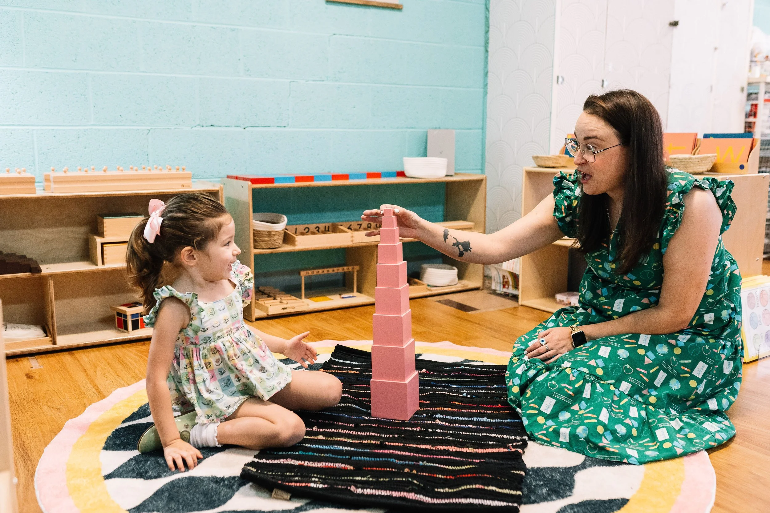 A young girl sitting on a colorful rug, playing Jenga with a woman in a classroom or playroom setting.