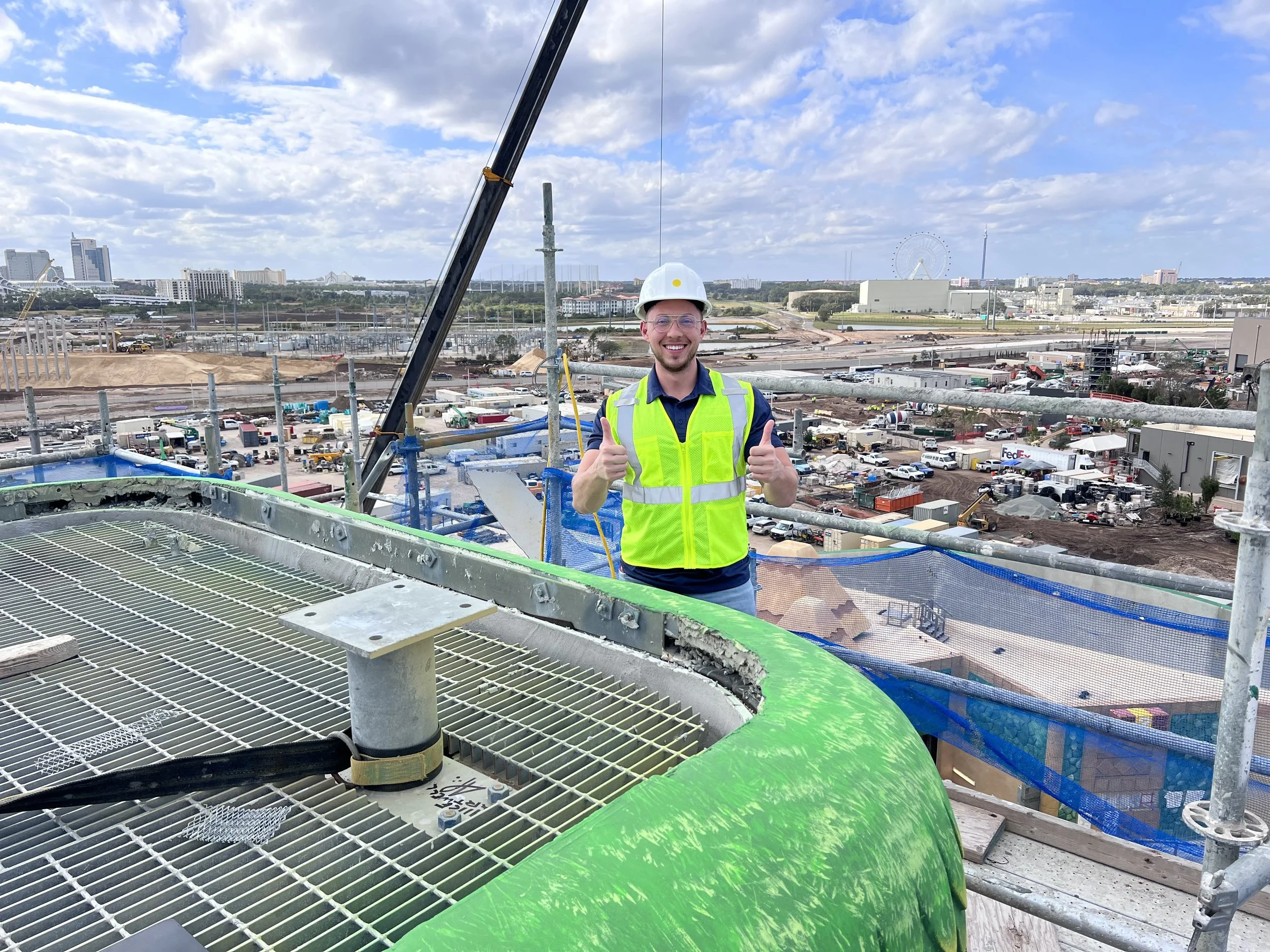 A smiling construction worker in a yellow safety vest and white hard hat giving a thumbs up on top of a building under construction with a cityscape and Ferris wheel in the background.