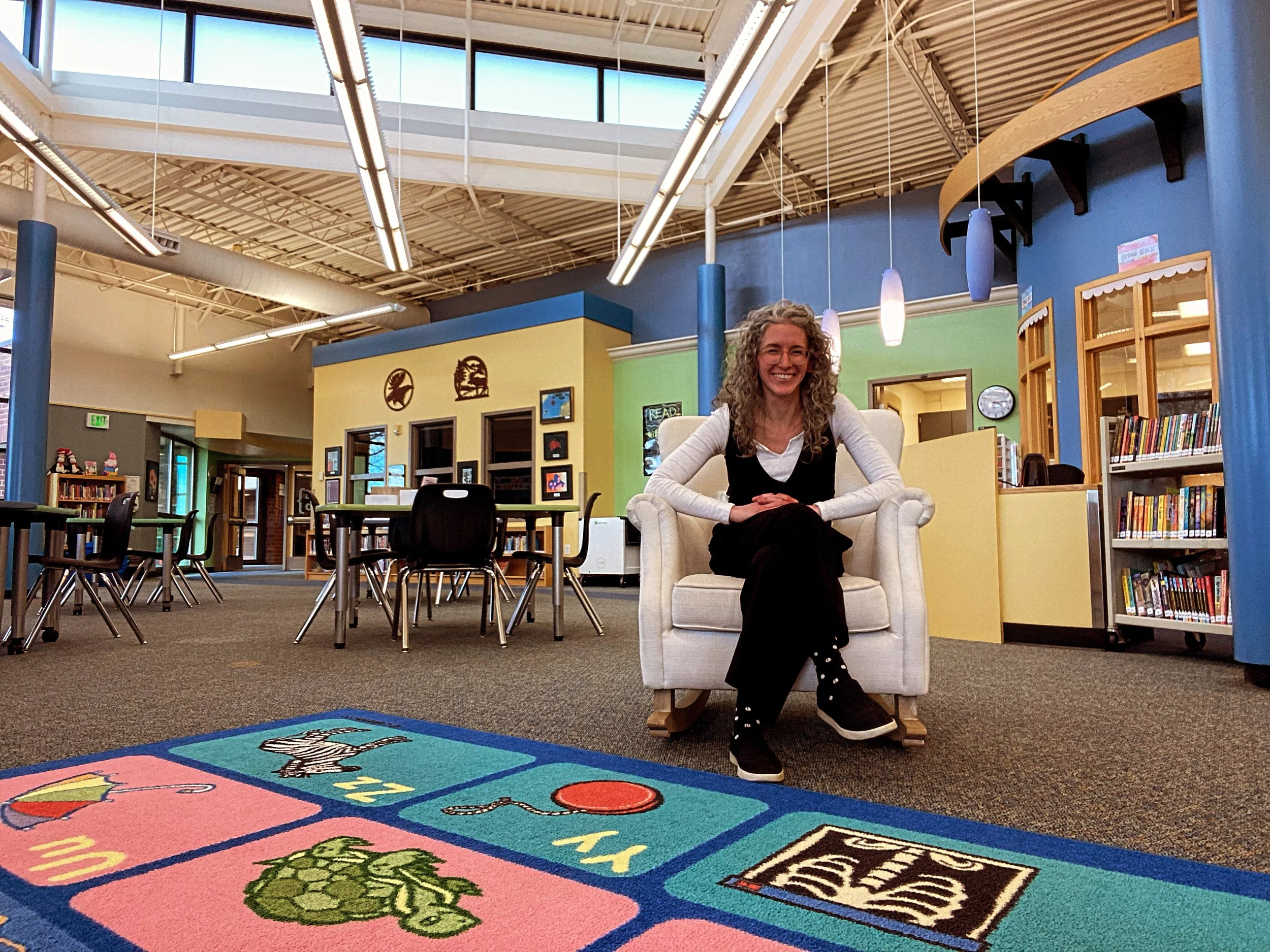 A smiling woman with curly hair, wearing glasses, a white long-sleeve shirt, and a black jumpsuit, sitting on a cream-colored armchair in a colorful library. There are tables, chairs, bookshelves, and decorative wall art in the background.