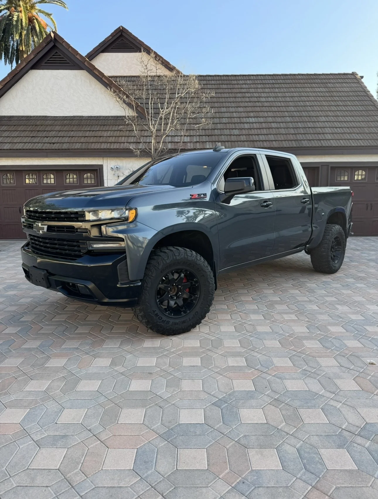 Gray four-door pickup truck parked on a hexagonal tile driveway in front of a house with a gabled roof.