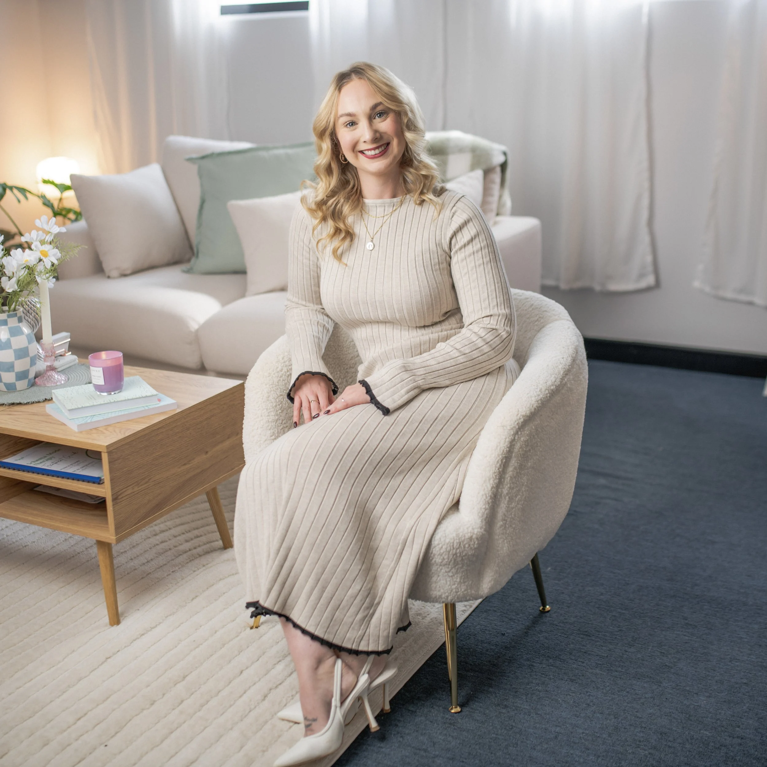 A woman with blonde curly hair, wearing a beige ribbed dress and white high heels, sitting on a cozy cream-colored chair in a modern living room, smiling.