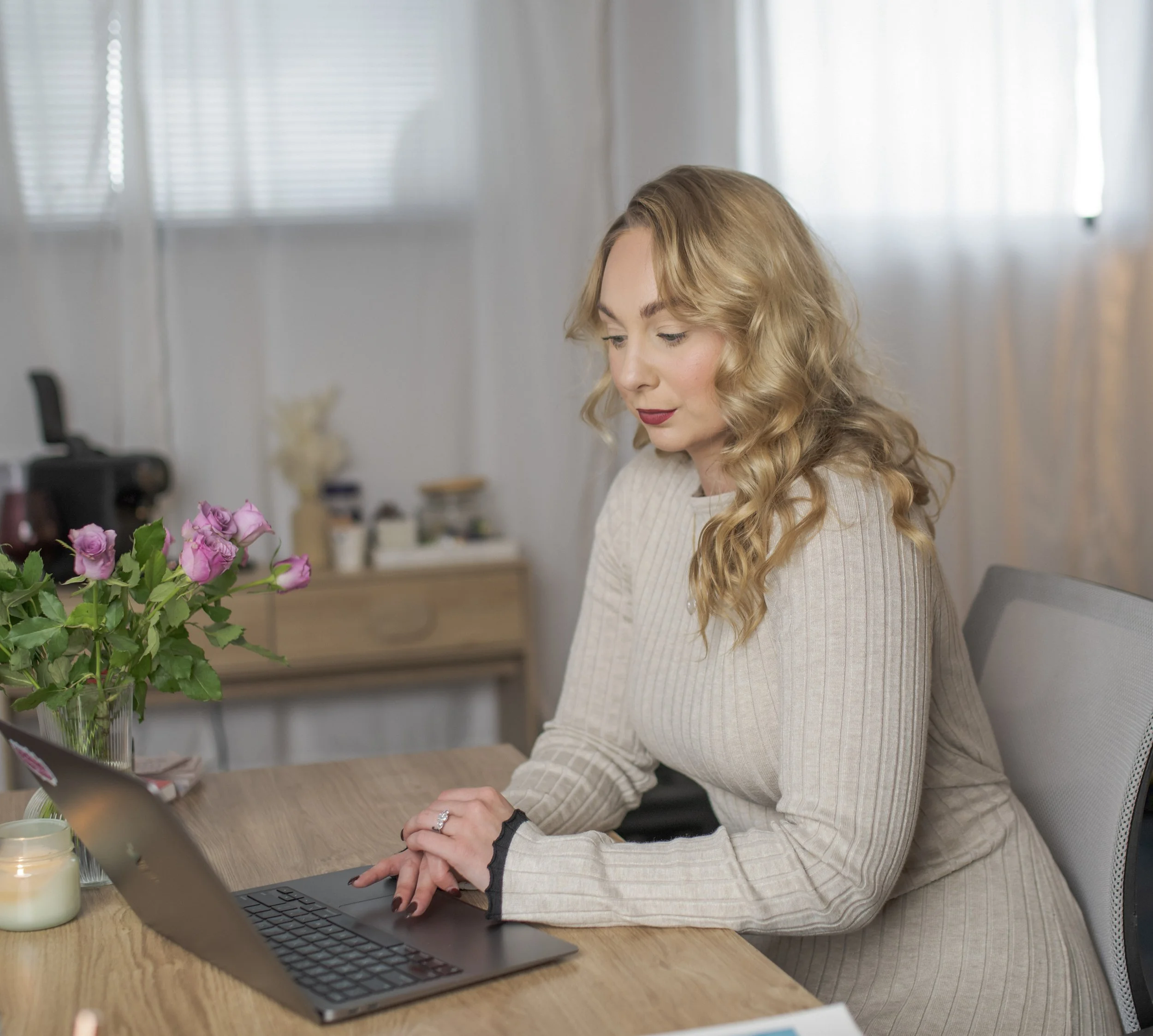 A woman with blonde curly hair sitting at a wooden table, working on a laptop. A vase with purple roses and a candle are on the table. The background shows a window with blinds, a small wooden drawer, and various items.