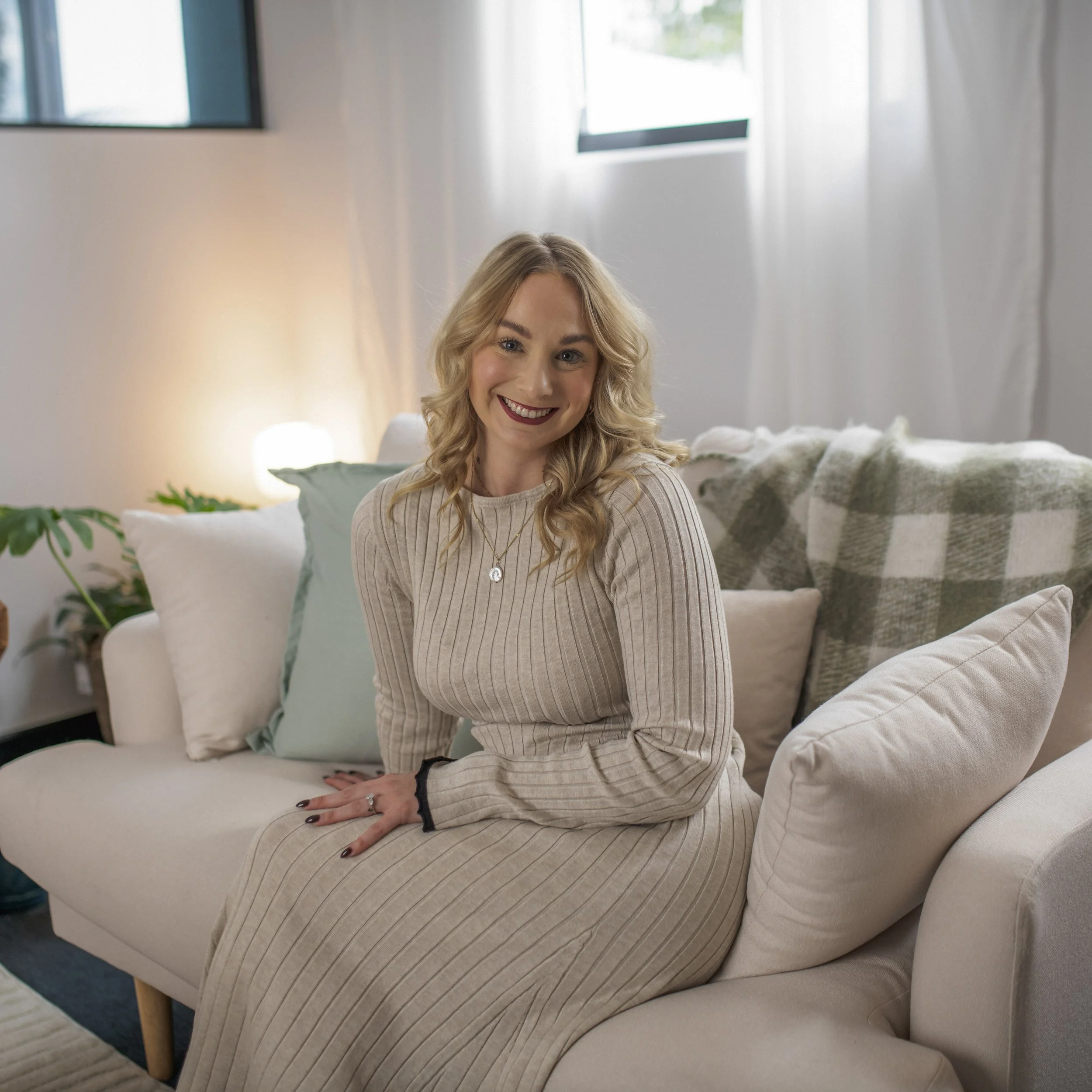 A woman with blonde curly hair sitting on a cream-colored couch in a cozy, well-lit living room, smiling at the camera.