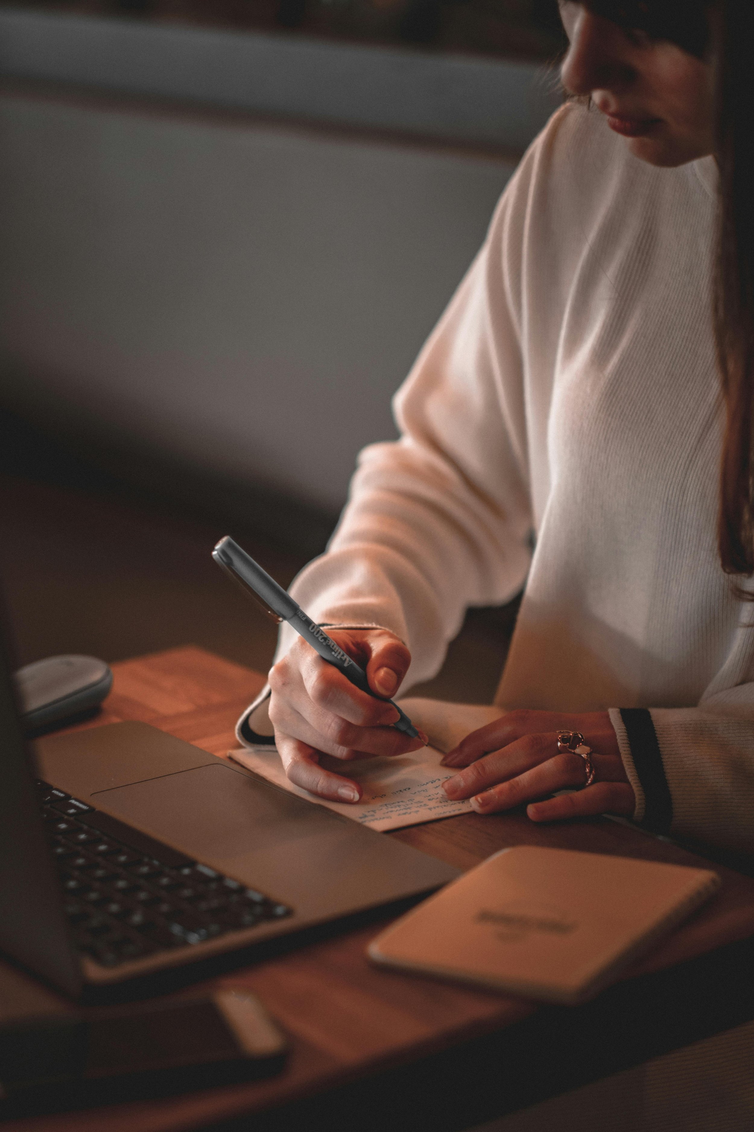 A woman writing notes on a small notepad at a wooden desk with a laptop and a smartphone nearby.