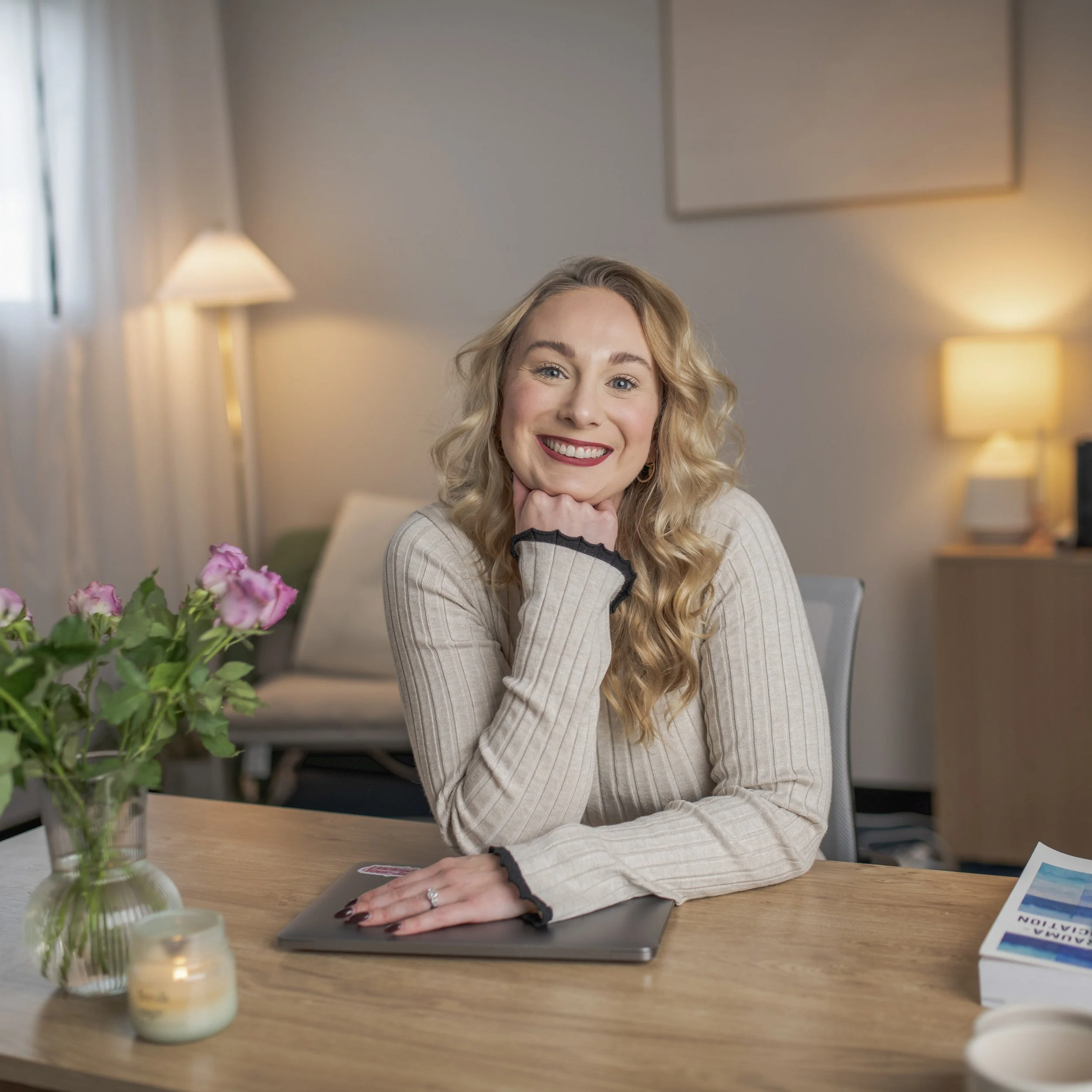 A woman with long blonde curly hair, smiling and resting her chin on her hand, sitting at a wooden desk in a cozy, softly lit room with a beige sweater, a laptop, a small vase with pink roses, a lit candle, and a book on the desk.