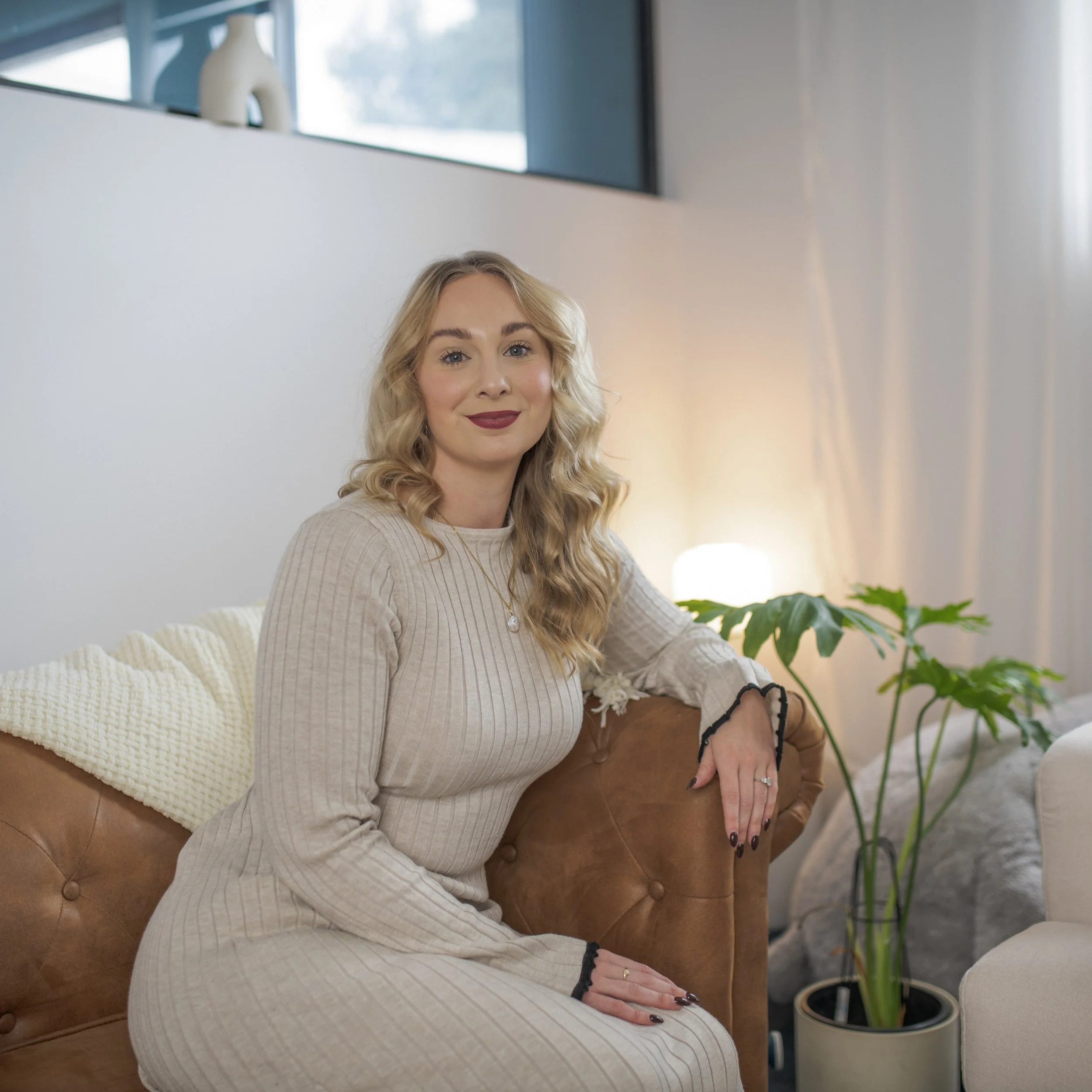 A woman with blonde curly hair sitting on a brown leather sofa in a modern living room, smiling at the camera, with a potted plant beside her and a warm light in the background.
