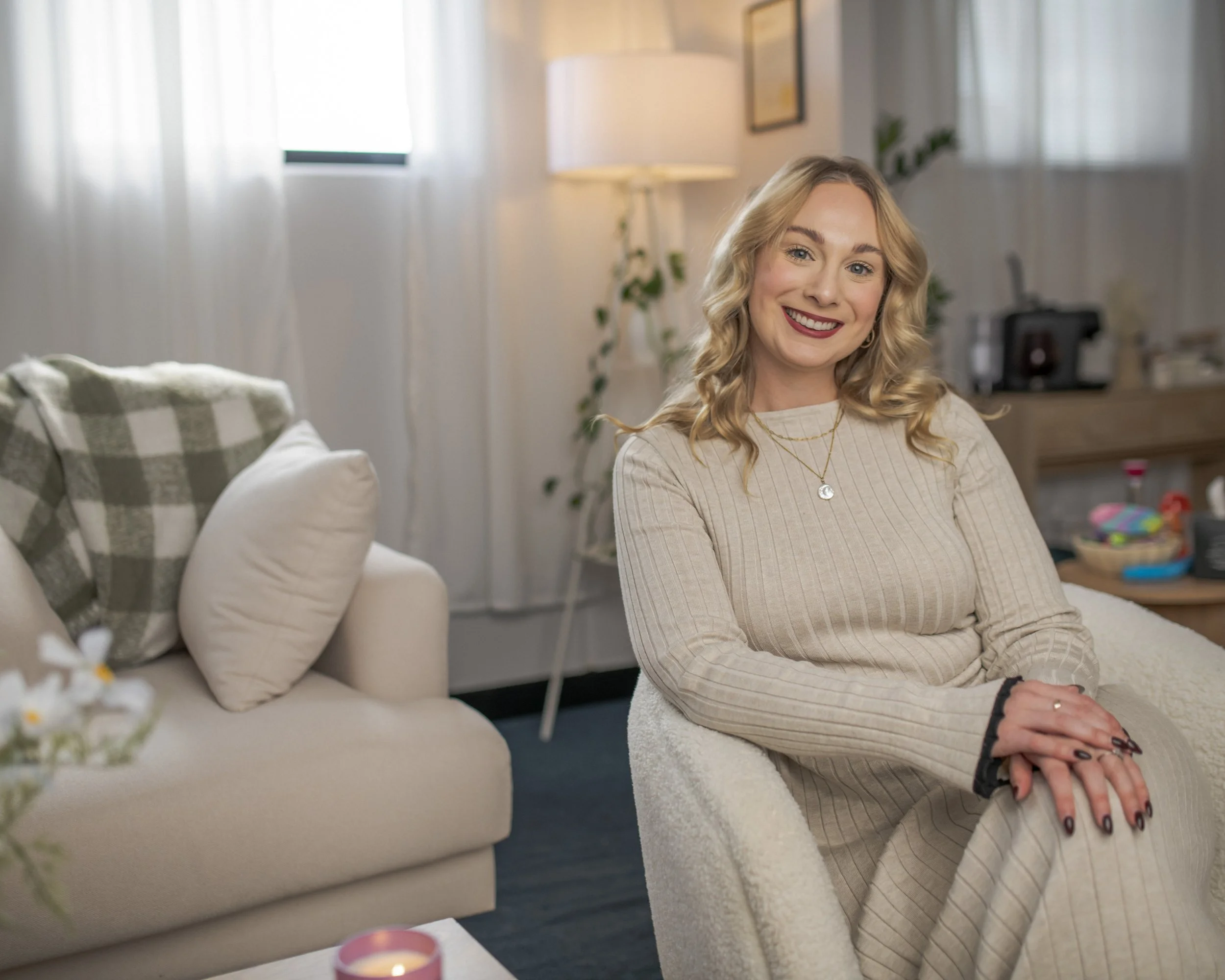 A woman with blonde hair and red lipstick sitting on a cozy chair in a well-lit living room, smiling at the camera.