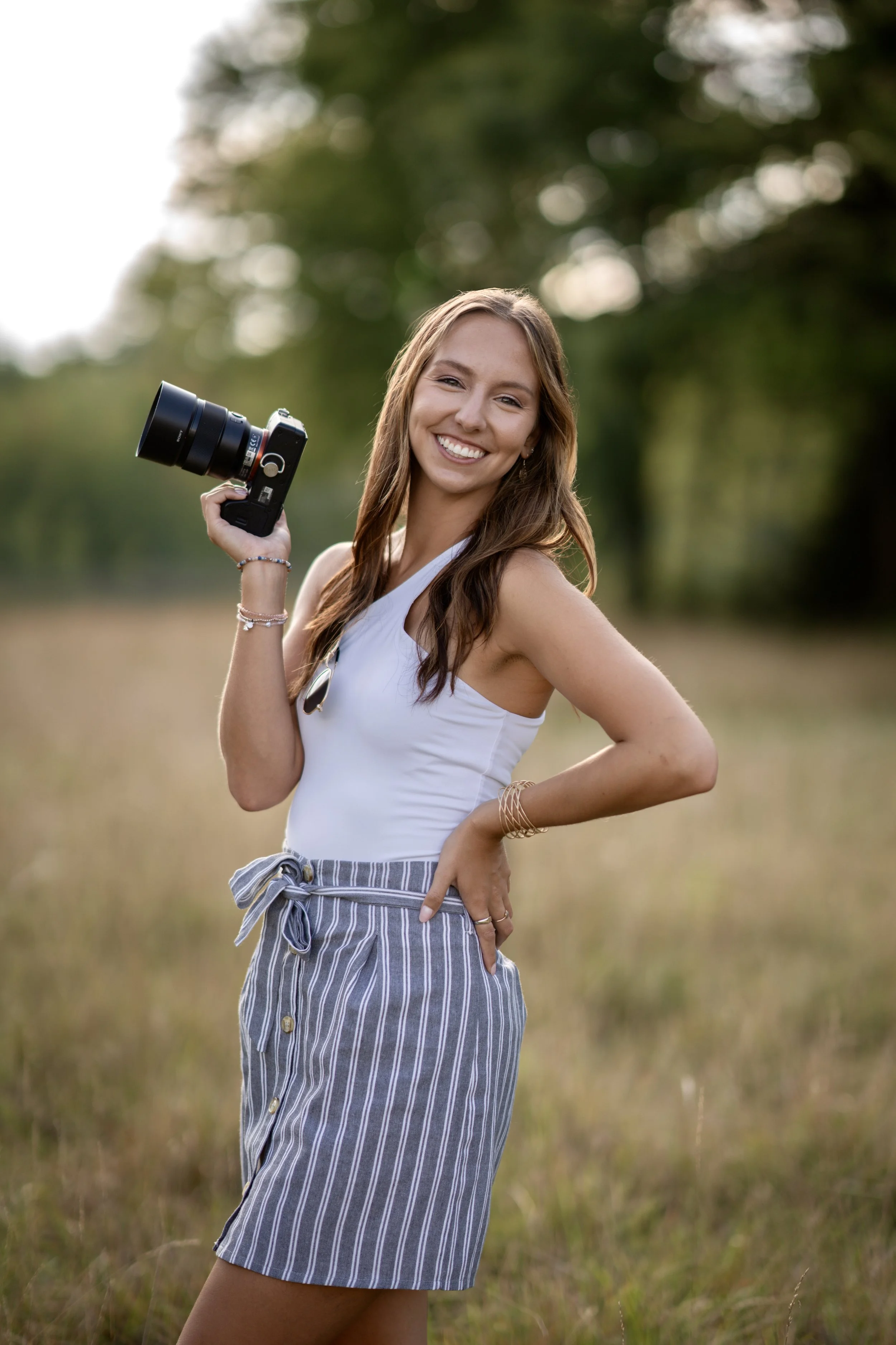 A young woman standing in a grassy field holding a camera, smiling at the camera, wearing a white sleeveless top and striped shorts, with trees in the background.