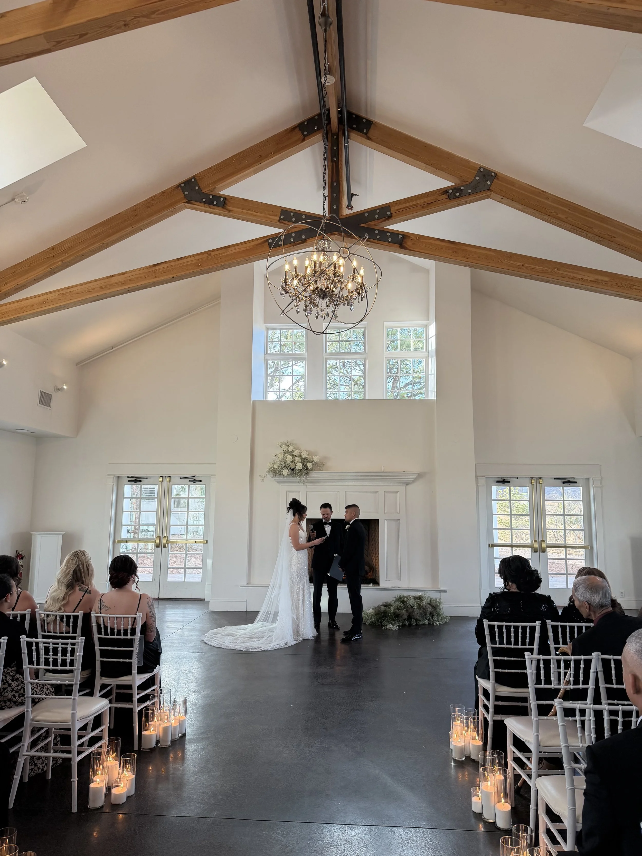 Indoor wedding ceremony with a bride and groom standing before officiant, guests seated on either side, candles on the floor, and a high ceiling with exposed wooden beams and a chandelier.