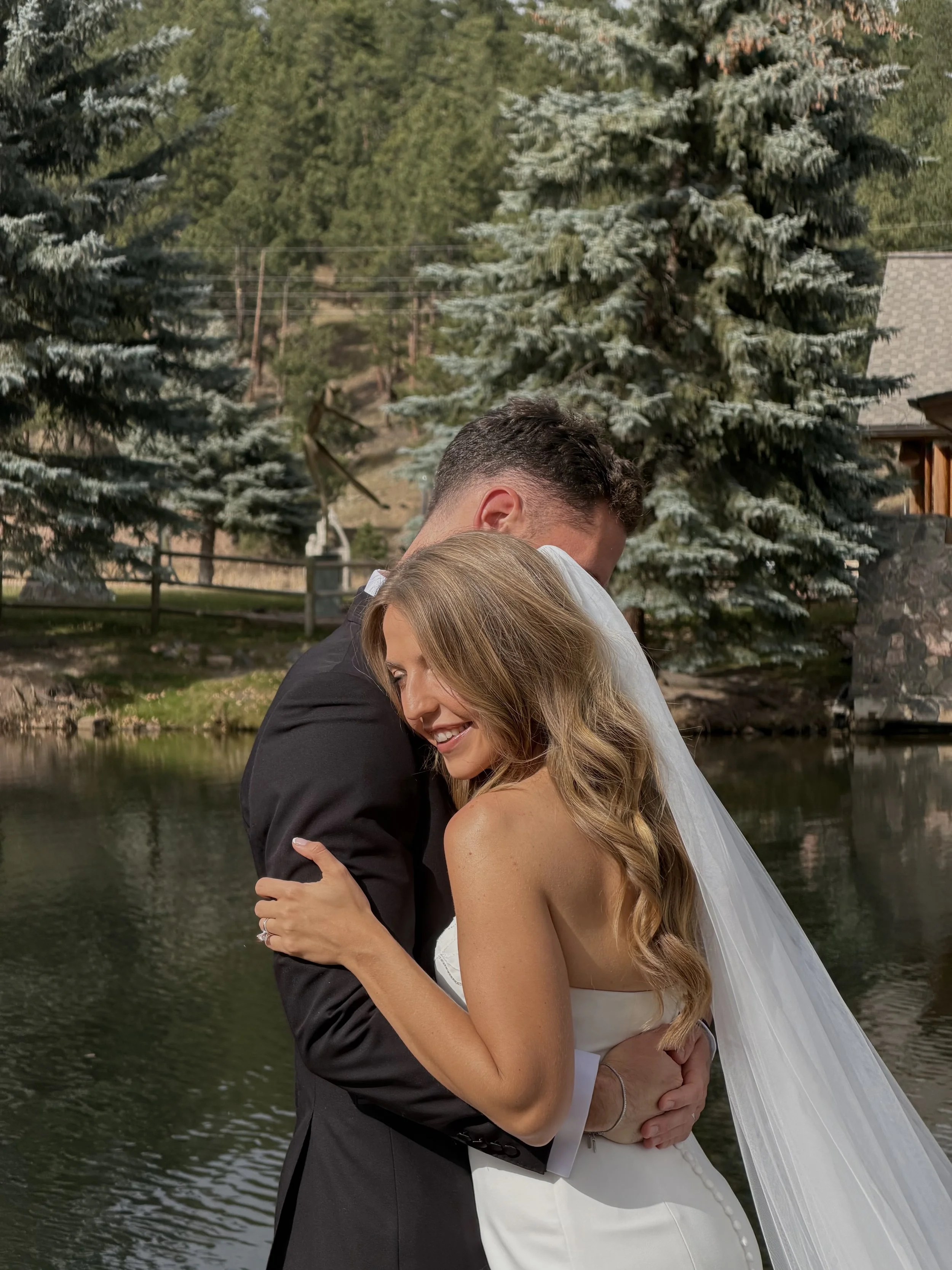 A bride and groom embracing outdoors near a pond, with trees and a house in the background.