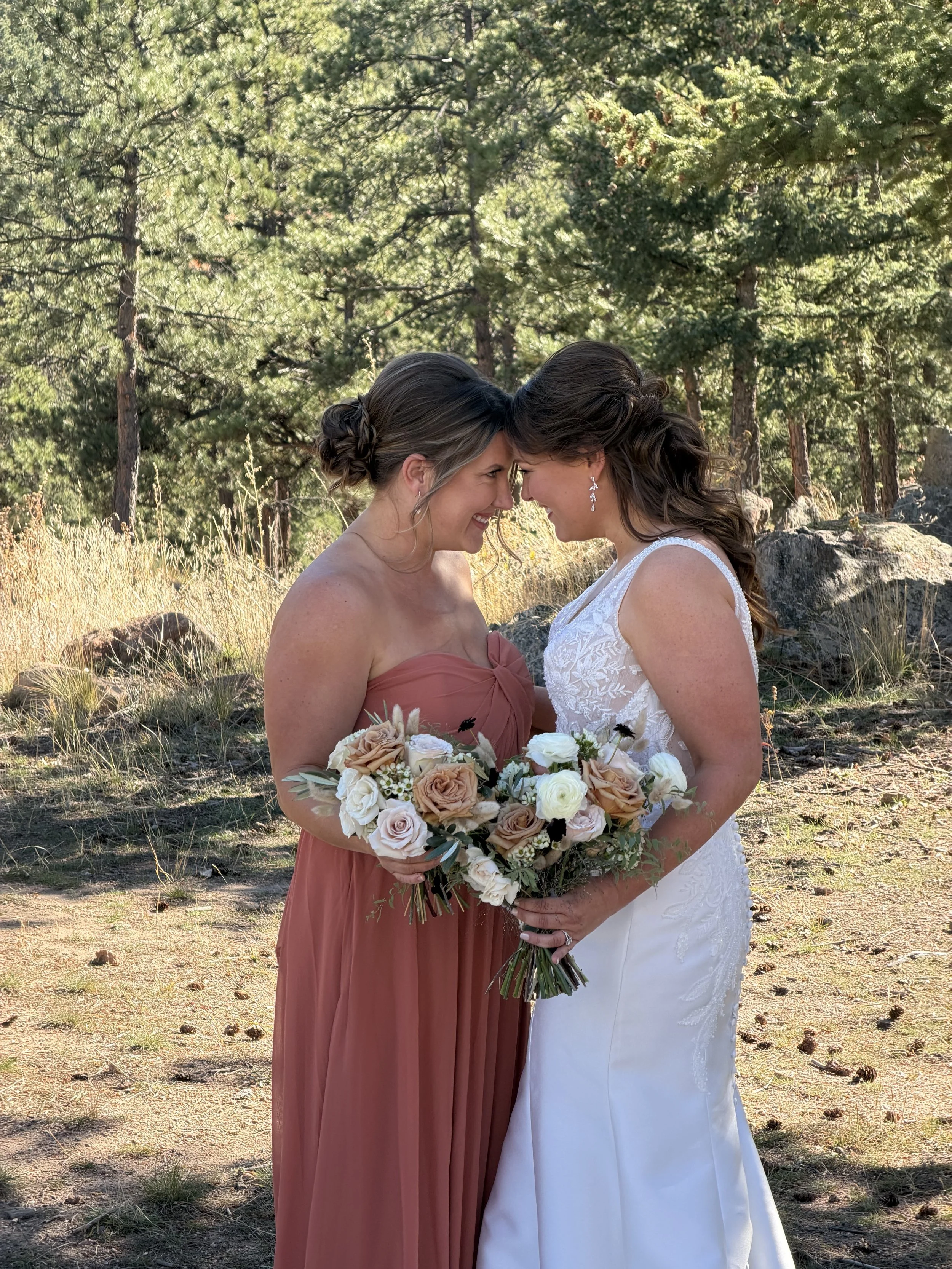 Two women, one in a white wedding dress and the other in a pink bridesmaid dress, are standing close, touching foreheads and smiling. They are holding a bouquet of flowers outdoors with trees in the background.