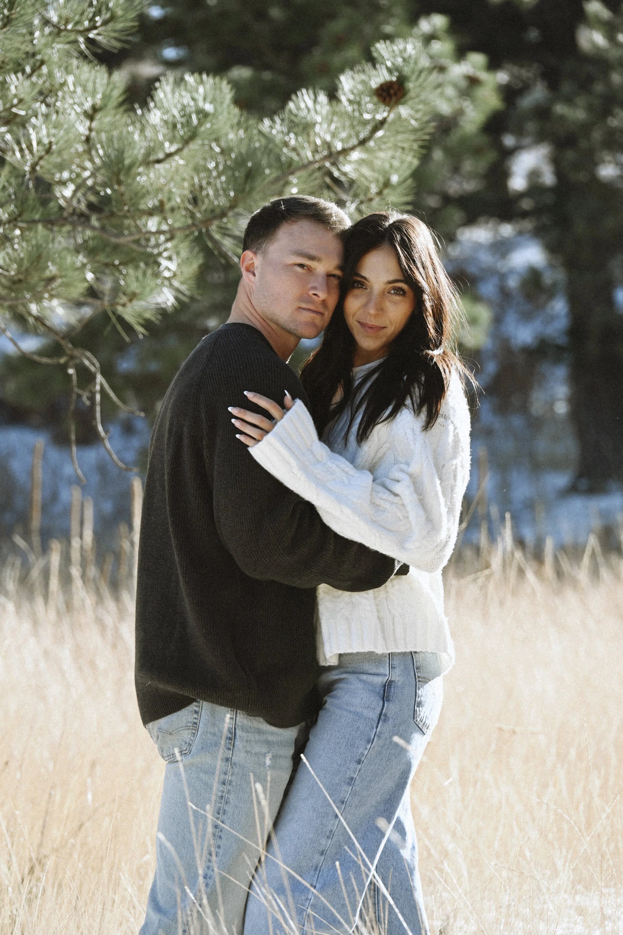 A young couple embracing outdoors in a field with tall grass, wearing sweaters, with trees in the background.