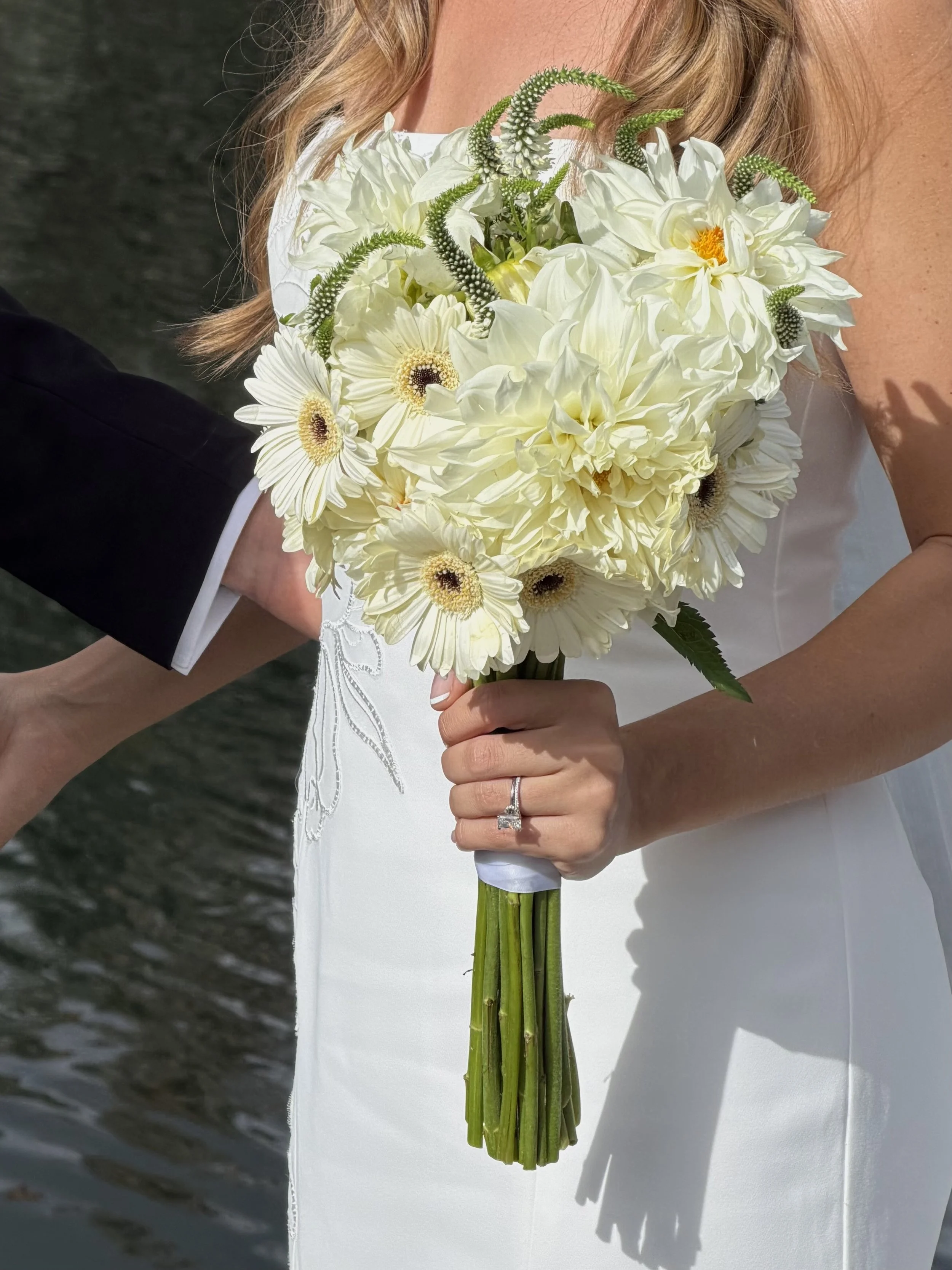 A woman in a white dress holding a bouquet of white flowers near water, with a ring on her finger.