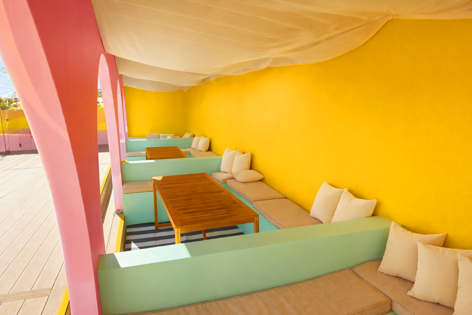 Colorful outdoor seating area with pastel green and pink walls, yellow background wall, wooden tables, and beige cushions and pillows under a white draped canopy.
