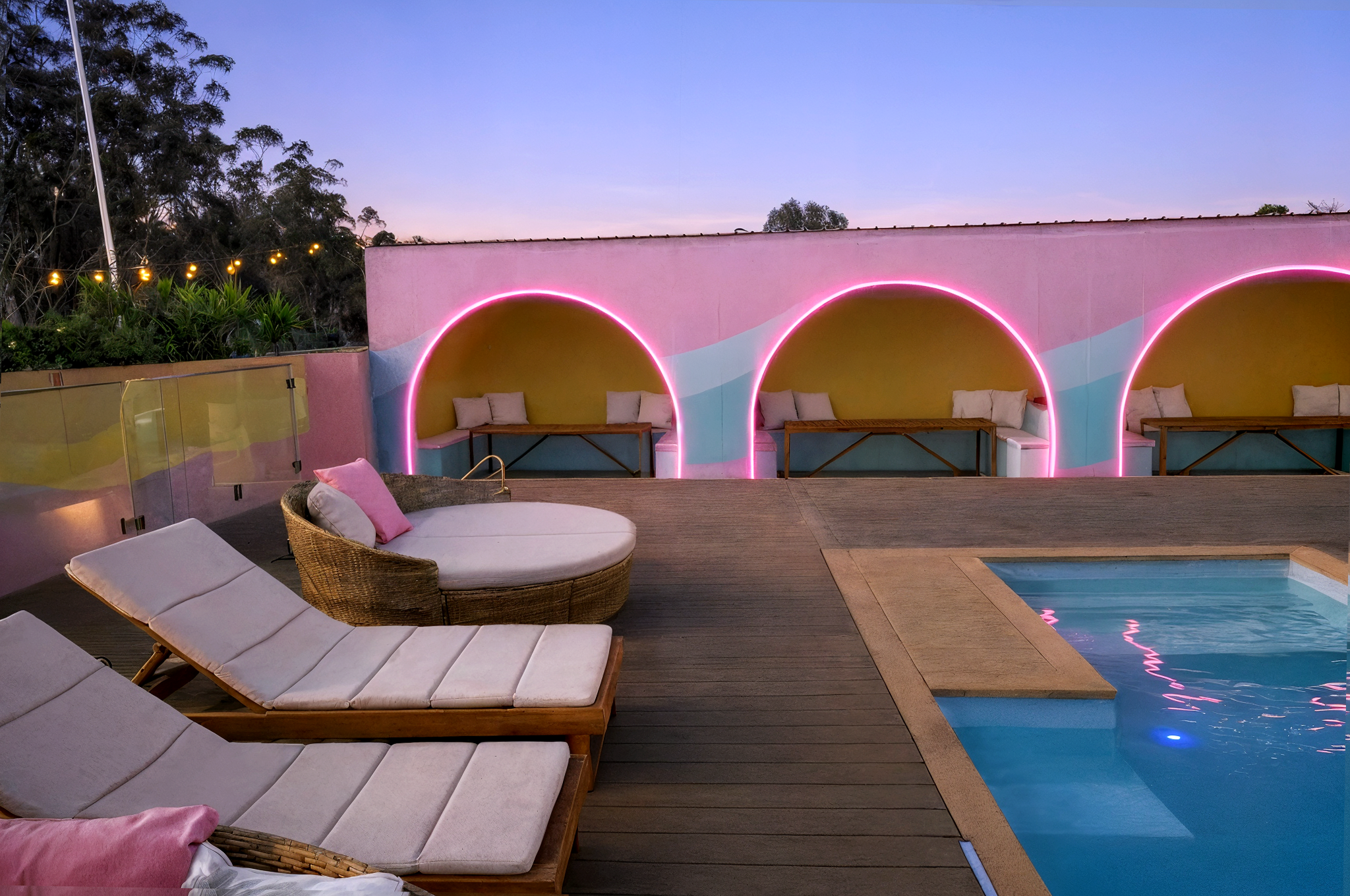 A rooftop pool area at dusk with lounge chairs, a round woven daybed, and decorative archways with pink neon lighting