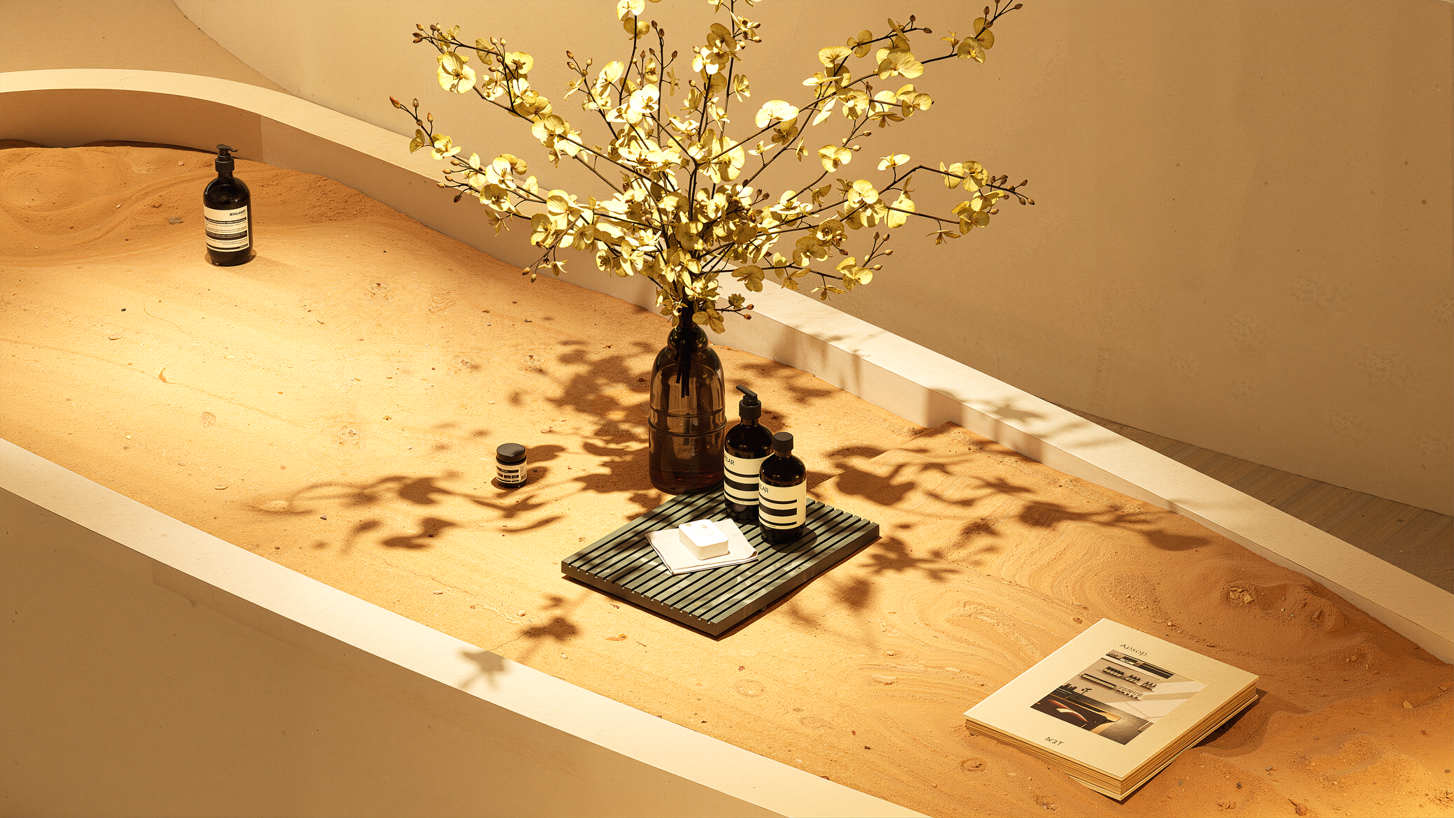 Decorative setup with a large brown vase holding a flowering branch, accompanied by various black and white bottles, a soap dish with a bar of soap, and a stack of papers or magazines on a sandy-colored surface.