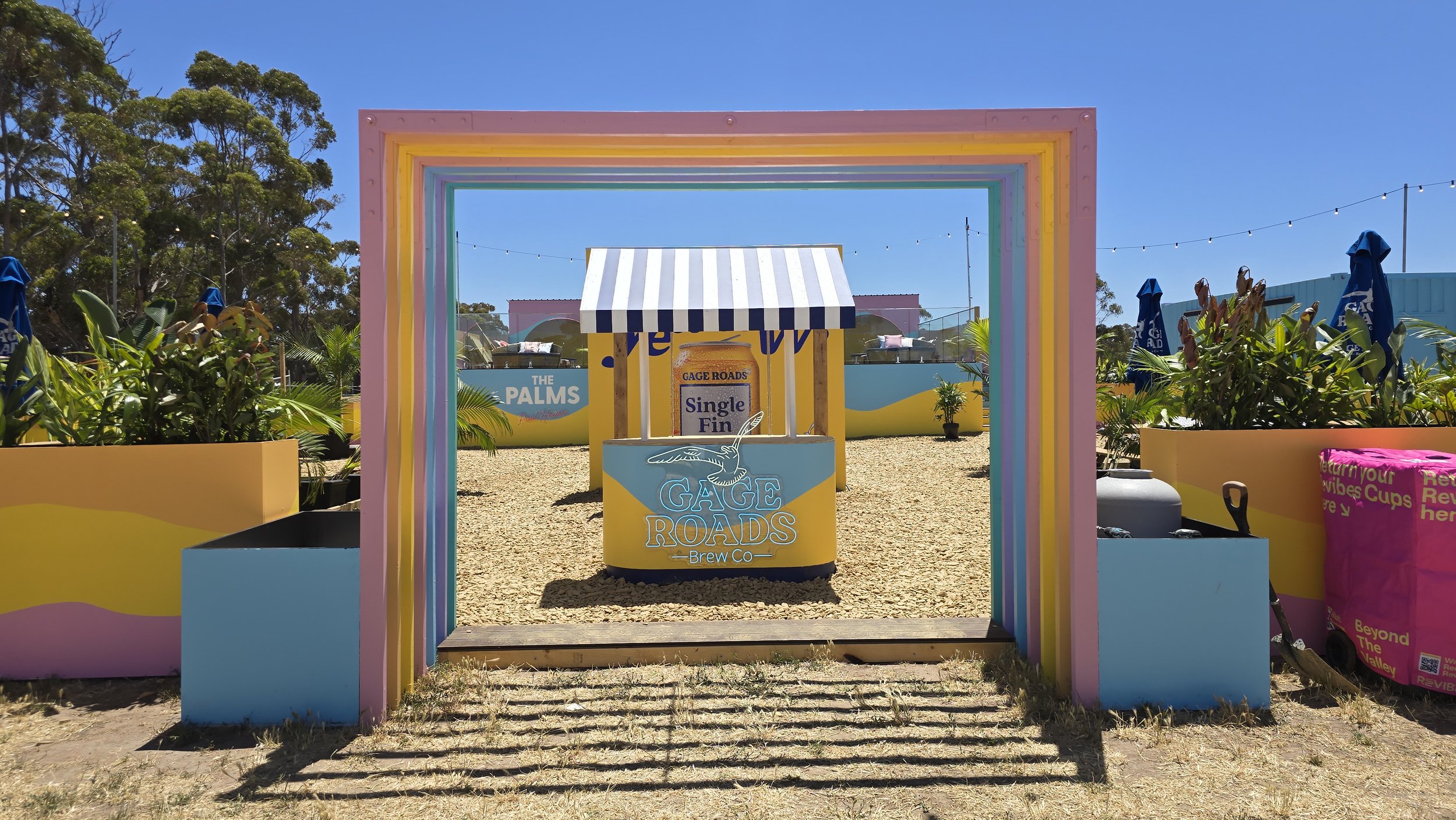 Colorful outdoor scene with a rainbow-colored frame leading to a small yellow stand with a blue sign for Gage Roads Brew Co, featuring a fish logo. Surrounding green plants, bright blue sky, and distance signs for The Palms and other attractions