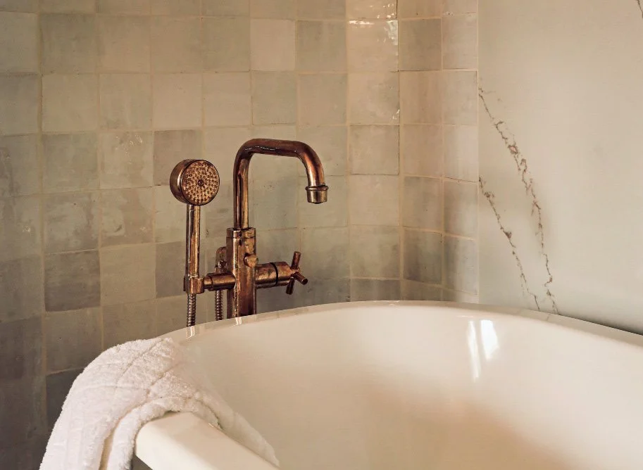 A vintage-style copper bathtub faucet with a handheld showerhead, mounted on a tiled bathroom wall next to a white bathtub with a towel draped over the side.