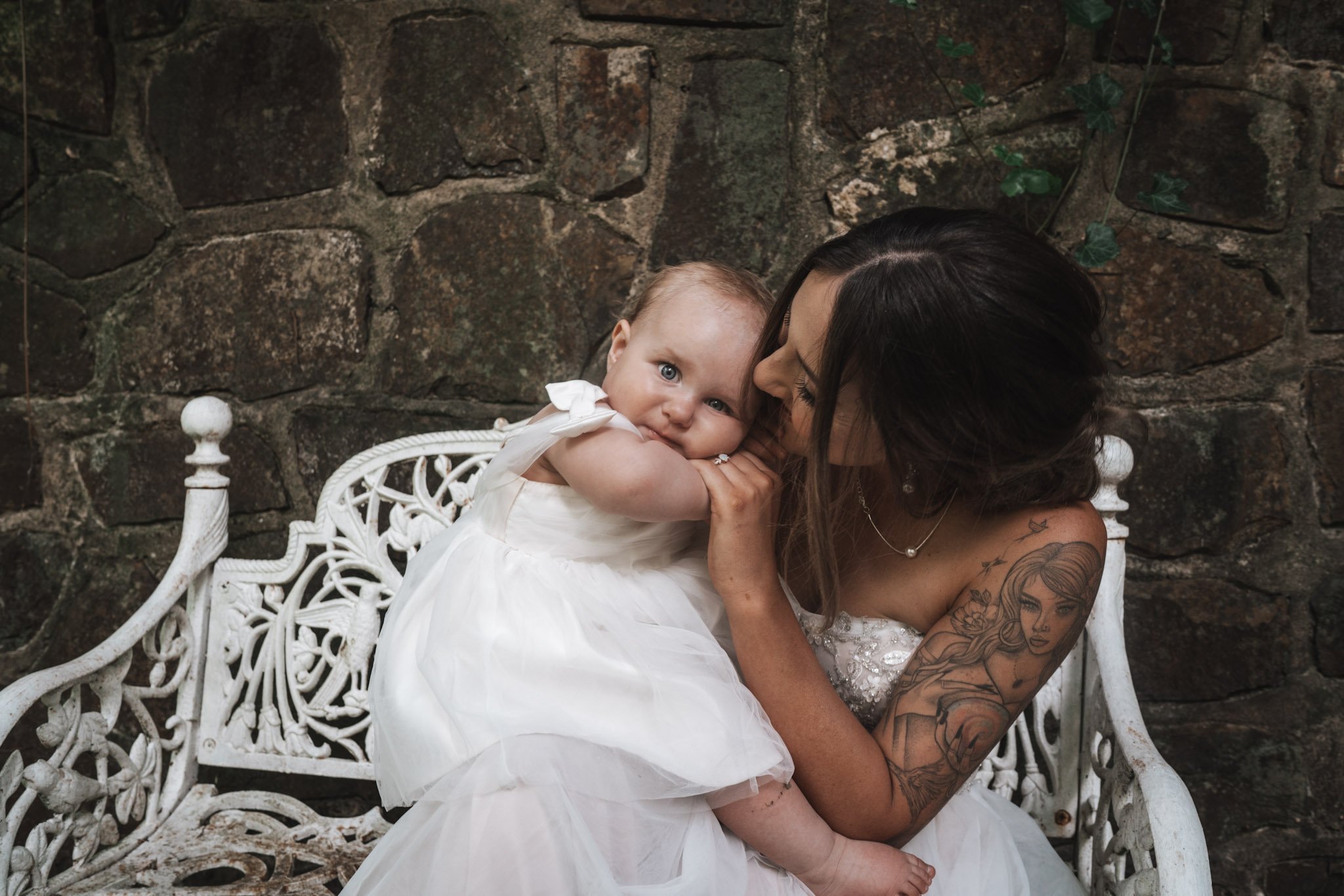 A woman with dark hair and tattoos on her arm, wearing a white dress, holding and hugging a young girl in a white dress with bows, sitting on a white ornate bench against a stone wall background.