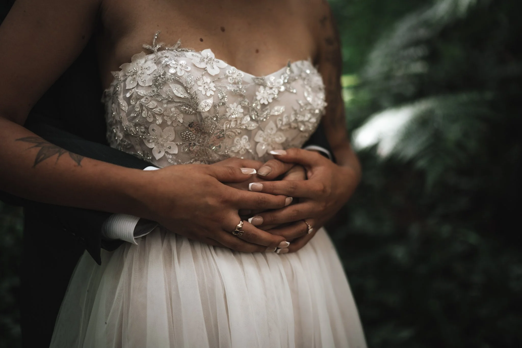 Close-up of a woman in a wedding dress with floral embroidery, holding her hands over her stomach, with a person behind her embracing her.