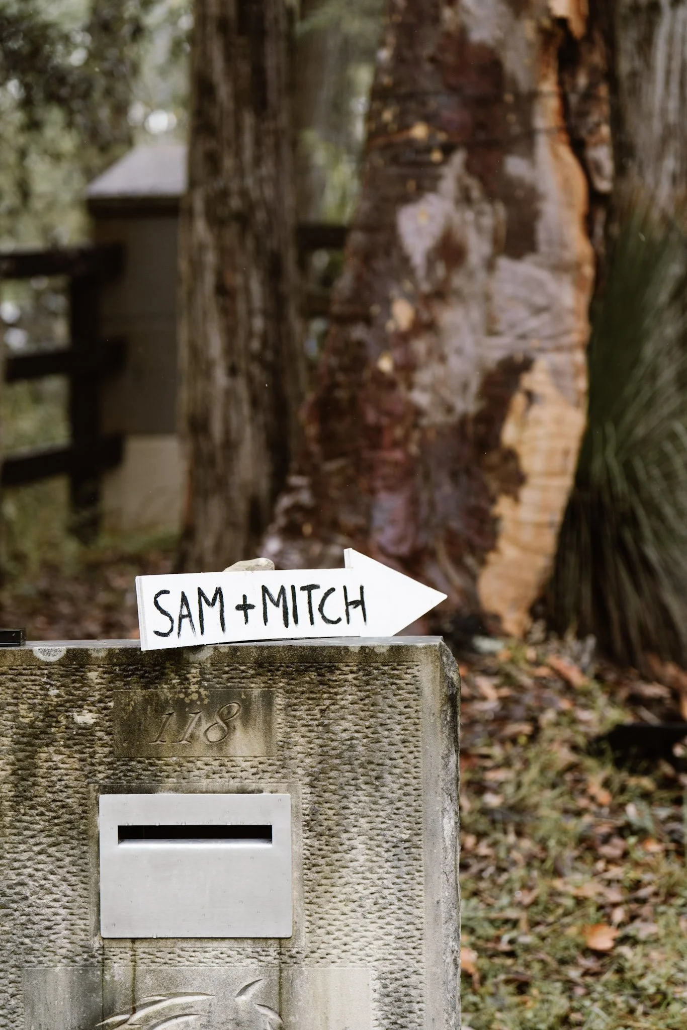 Sign on a mailbox with the text 'SAM + MITCH' and an arrow pointing to the right, with trees and a house in the background.