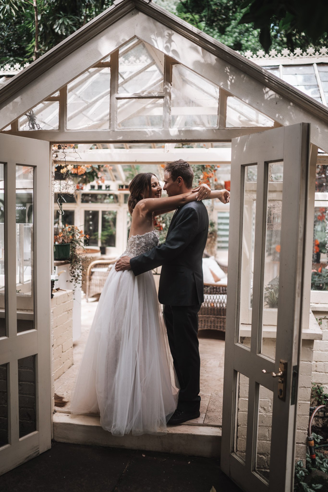 A bride and groom embrace inside a glass-roofed greenhouse, smiling at each other.