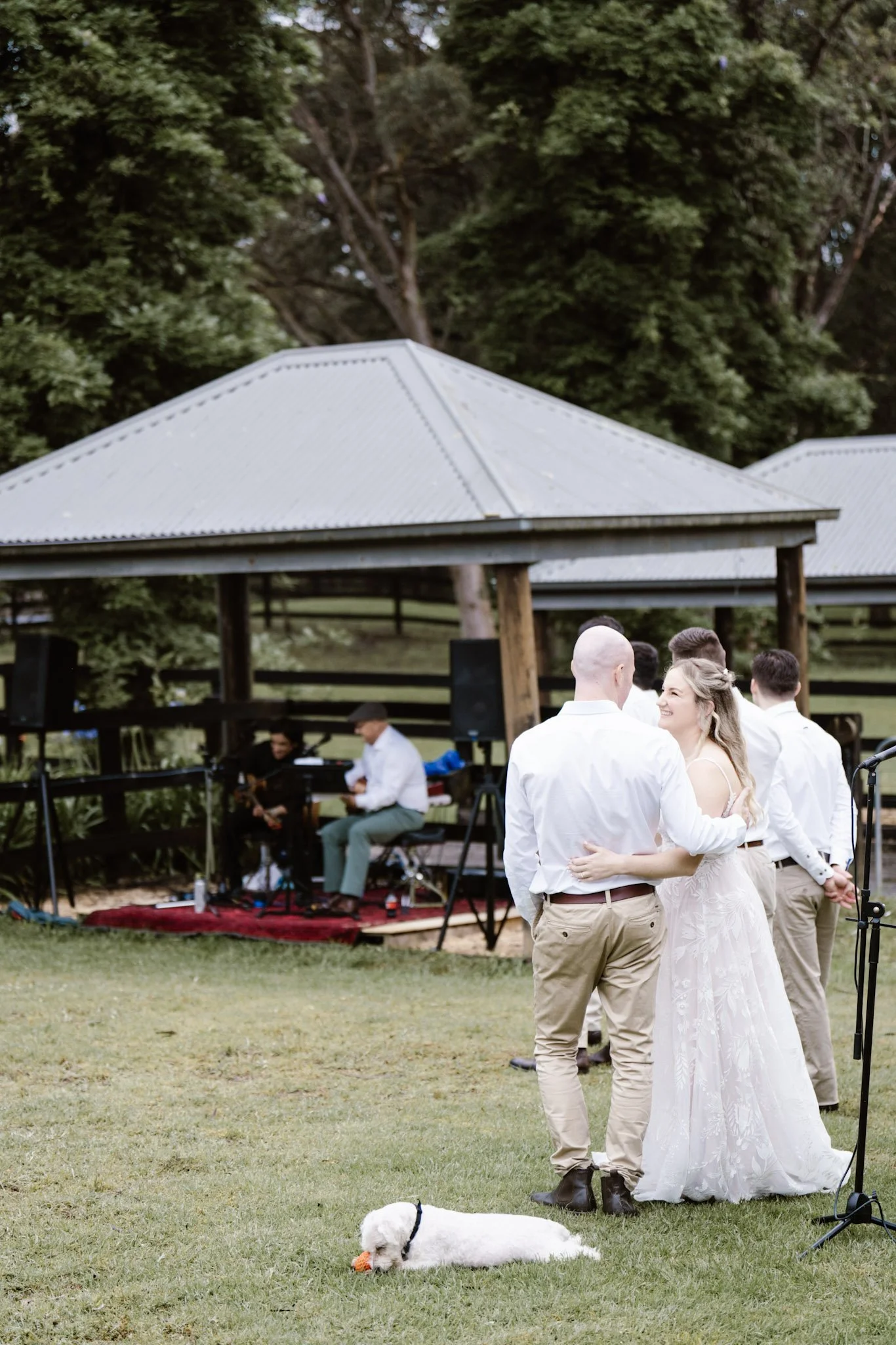 A wedding reception outdoors with a bride in a white gown and groom in beige pants dancing together, surrounded by other guests. There is a small dog lying on the grass. In the background, a band with two musicians is playing under a metal-roofed she