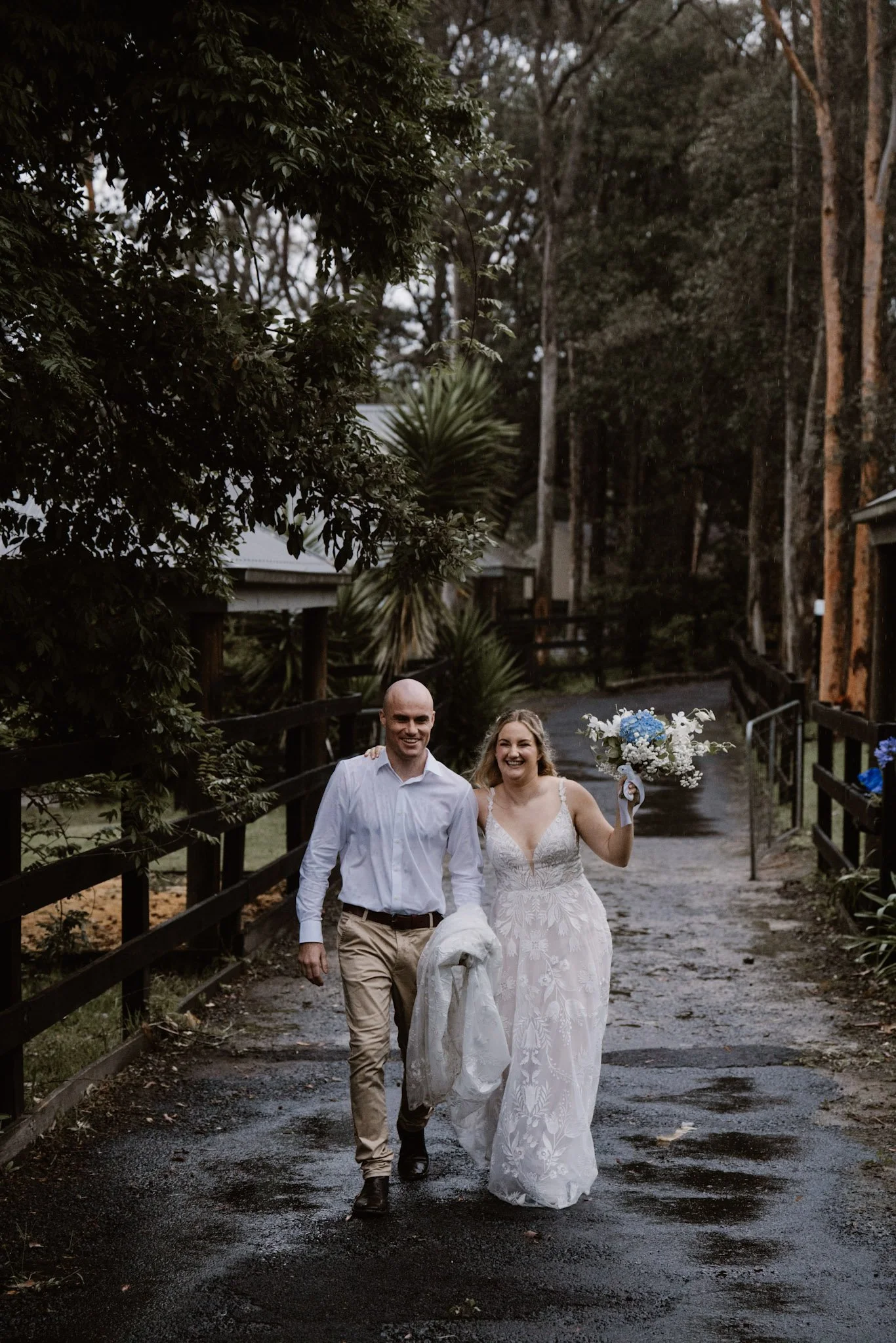 A newly married couple walking arm-in-arm outdoors on a wet, dark paved pathway surrounded by trees and greenery, with the woman holding a bouquet of white and blue flowers.