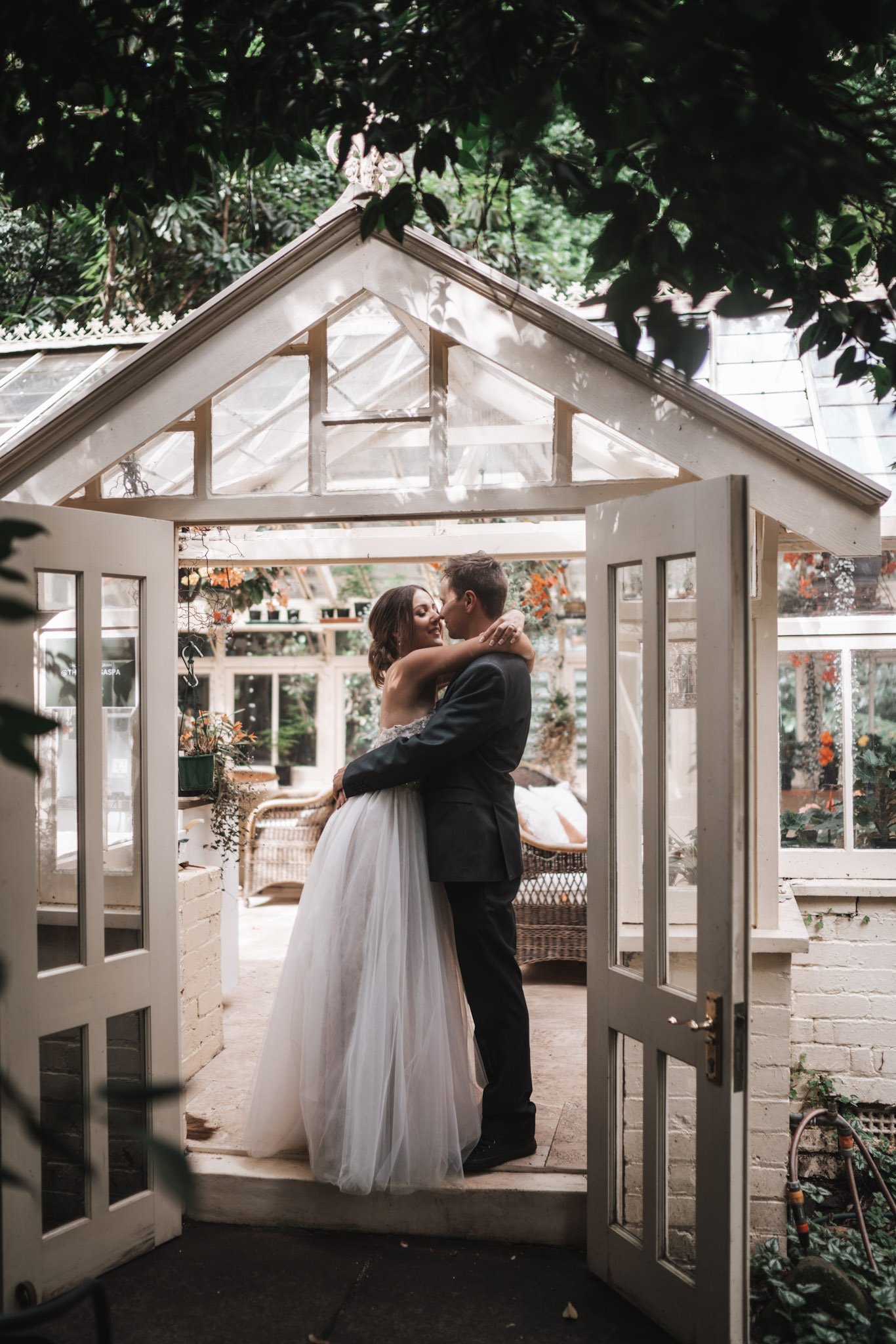A bride and groom sharing a kiss inside a glass-walled garden greenhouse.