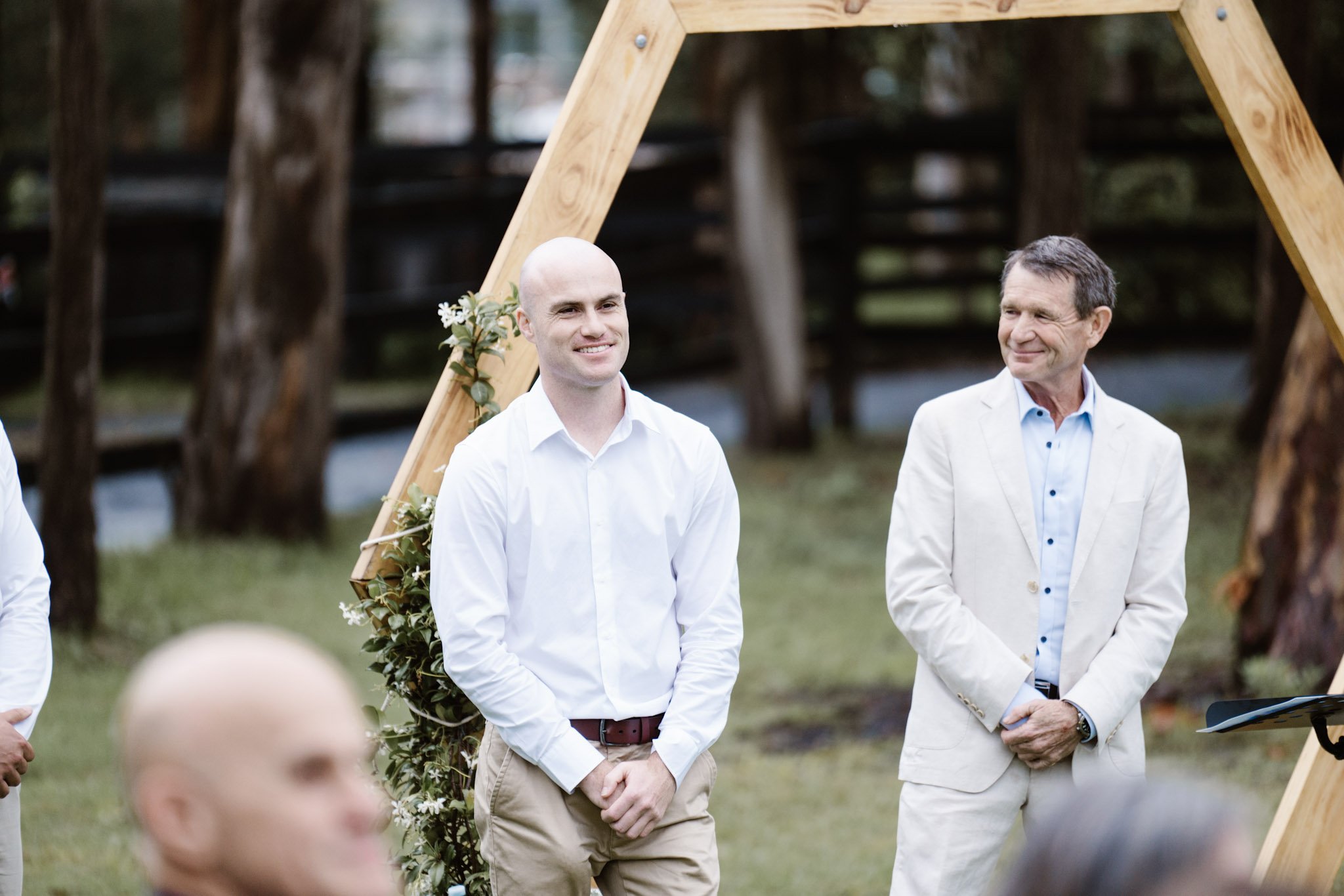 Two men standing outdoors, attending a wedding ceremony, with an arch decorated with flowers behind them.