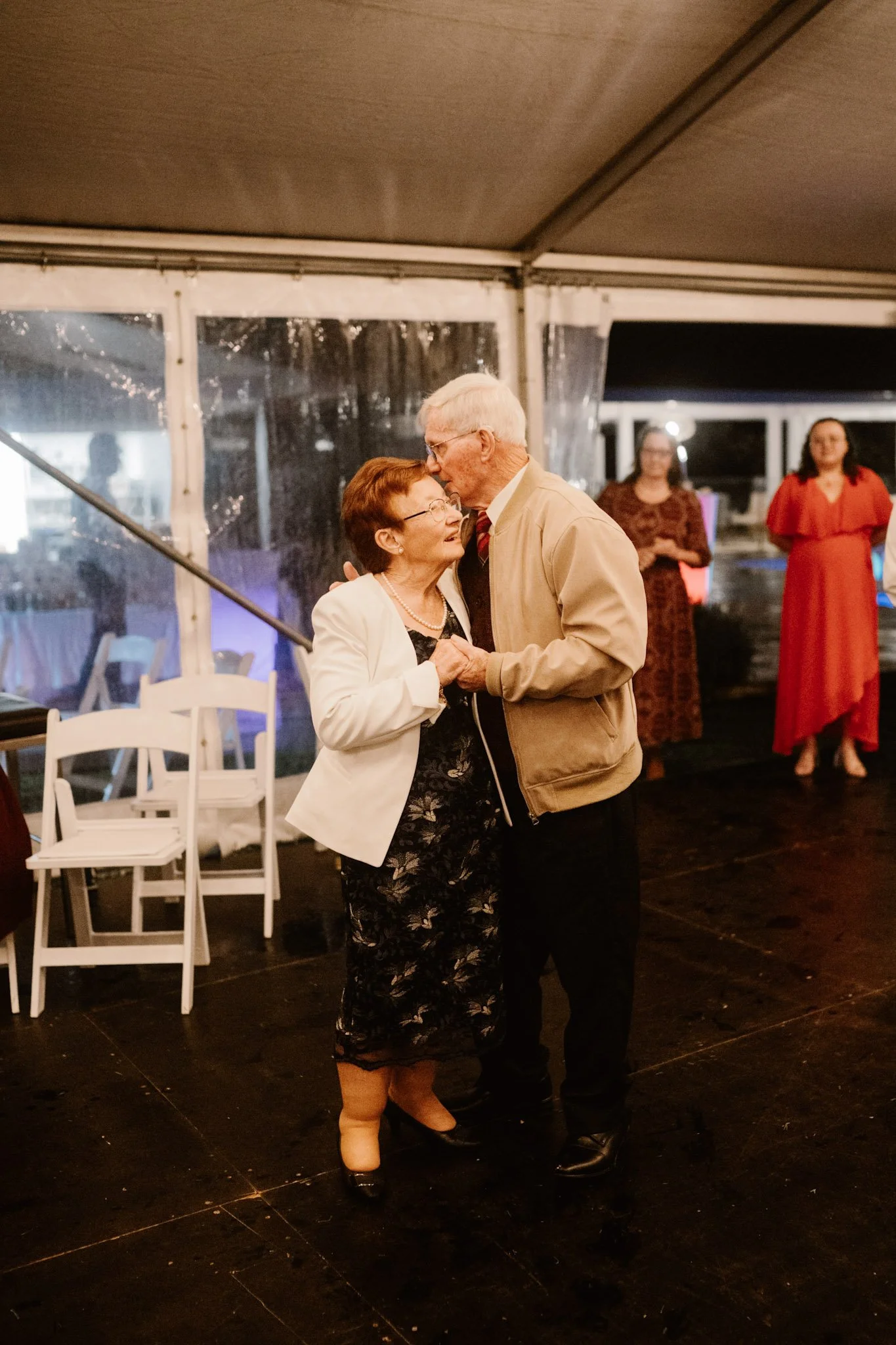 An elderly couple dancing together at a celebration inside a tent, with two women standing in the background, one in a red dress and the other in dark attire.