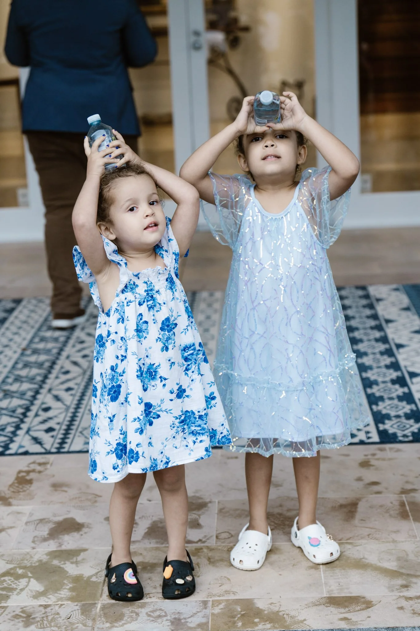 Two young girls in dresses holding water bottles on their heads, standing indoors on a wooden floor with a patterned rug in the background, and an adult partially visible behind them.