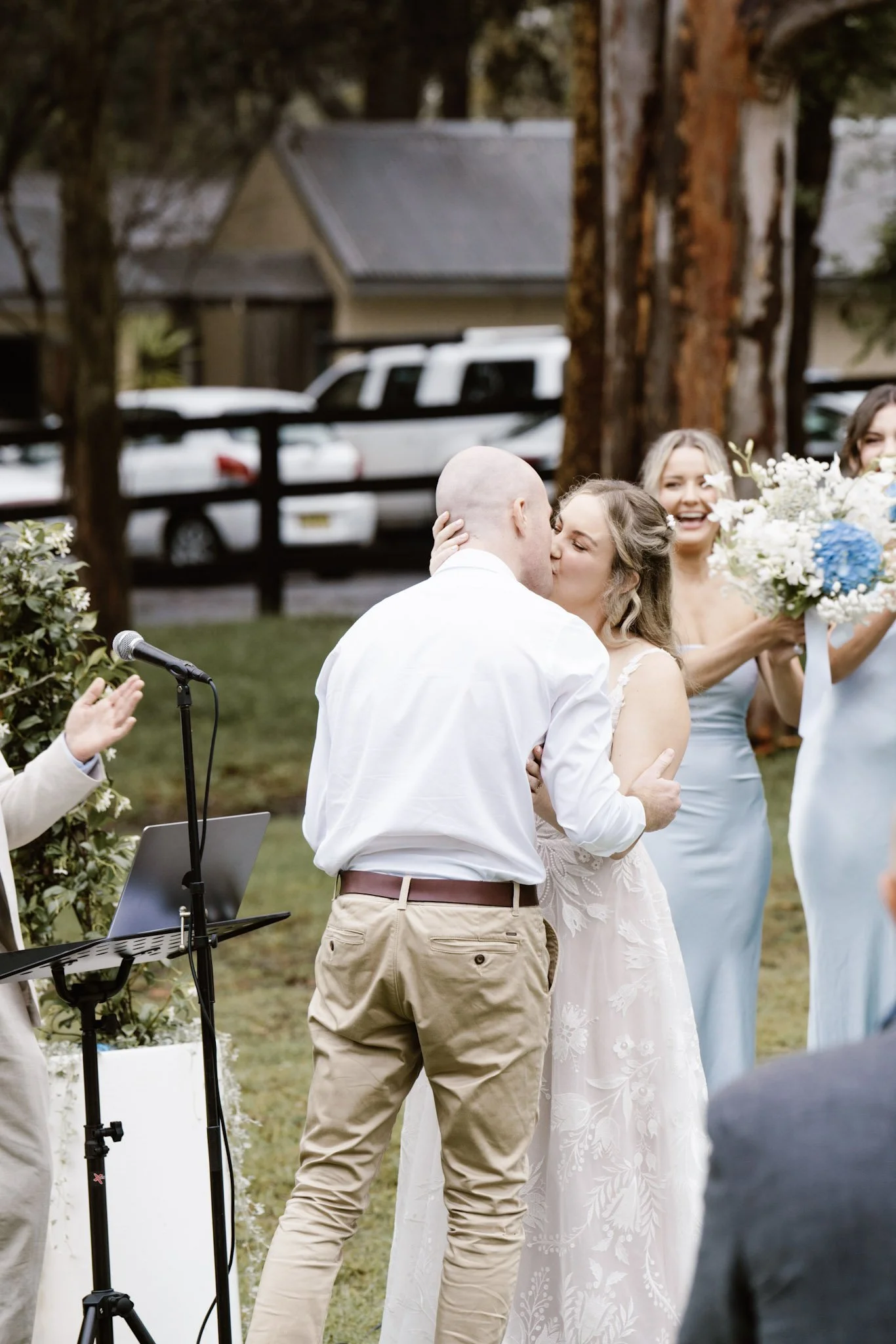 A newly married couple sharing a kiss during their outdoor wedding ceremony, with friends smiling and holding flowers nearby.