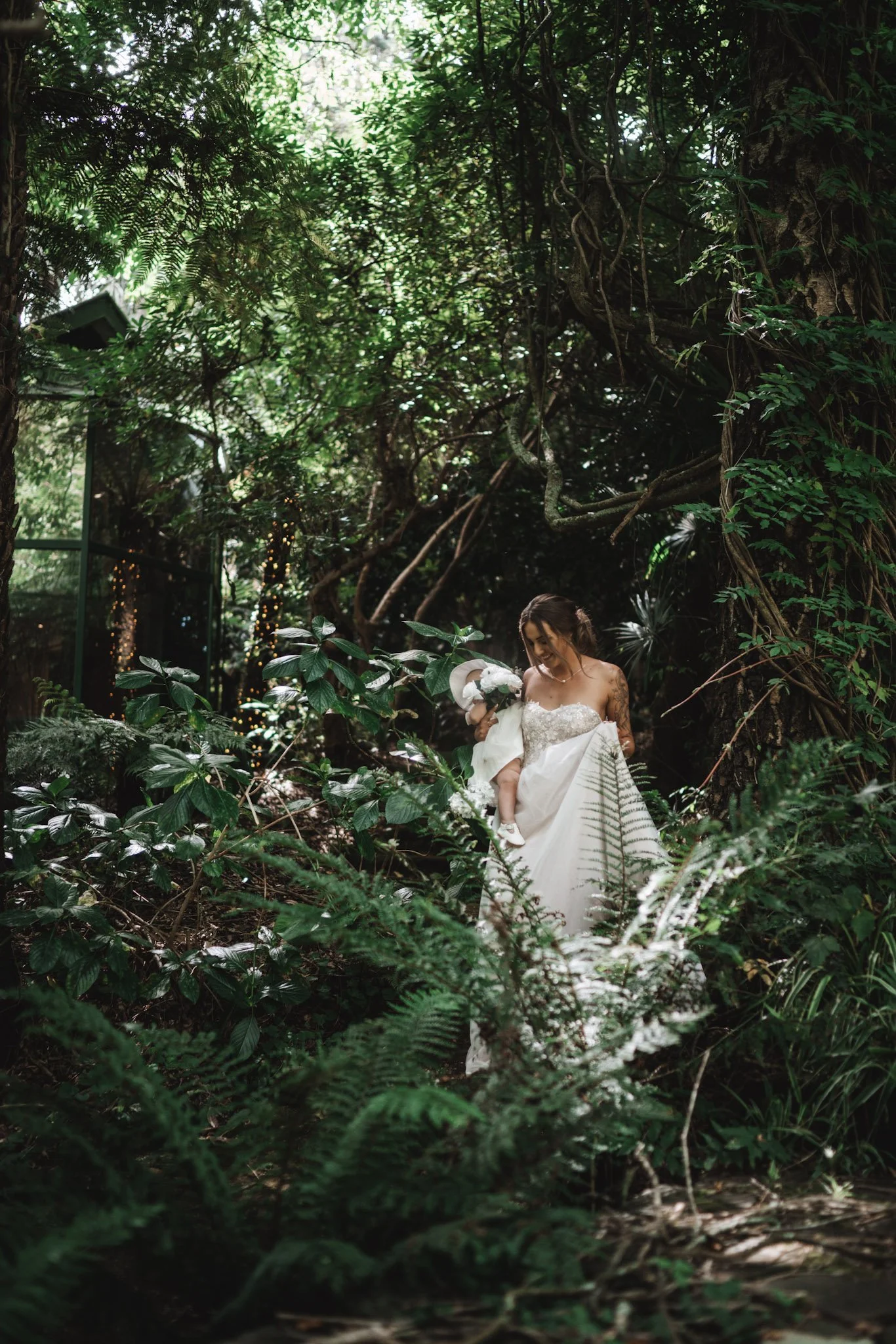 A bride in a wedding gown holding a young girl, surrounded by lush green plants in a forest setting.